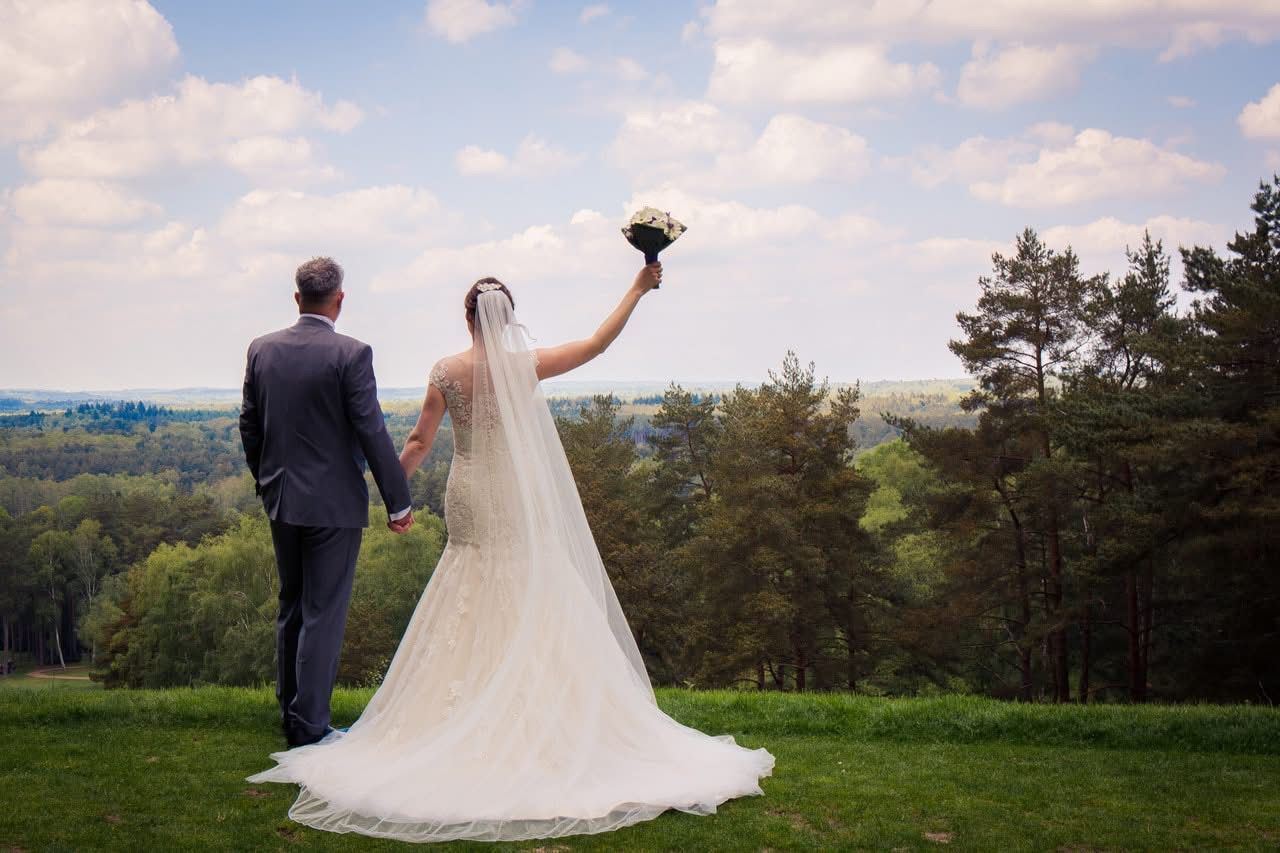 Bride and groom holding hands outdoors, bride raising bouquet, with scenic green landscape and blue sky behind them.