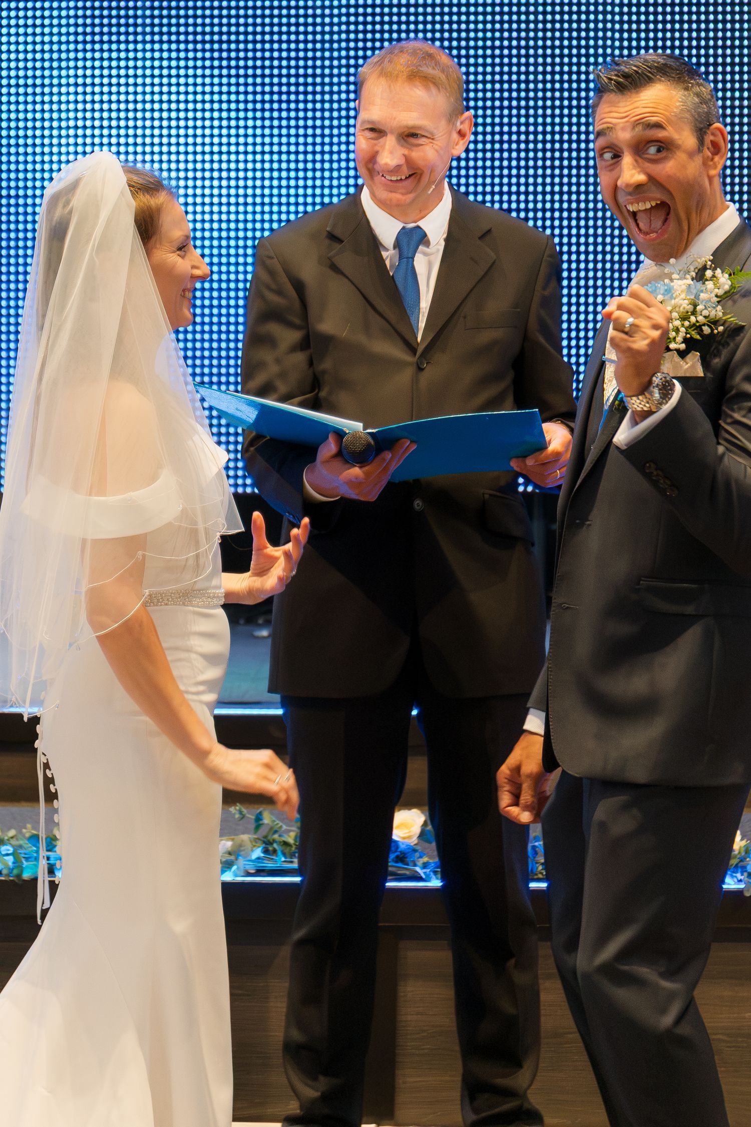 Joyful groom reacts with excitement during wedding ceremony as officiant reads vows to smiling bride in white gown and veil.