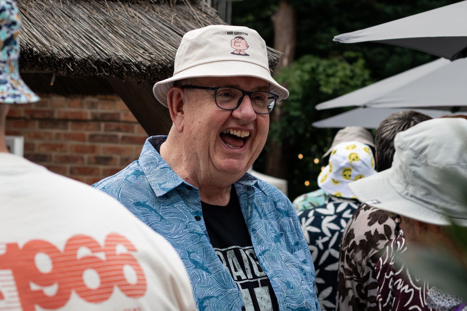 A laughing older man in a white fedora and blue shirt enjoys himself at an outdoor social gathering.