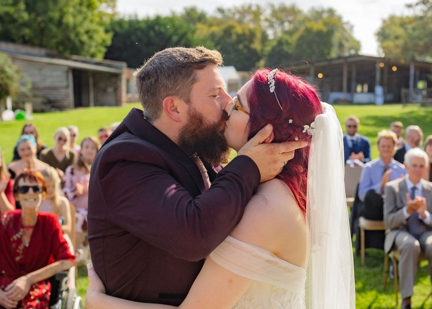 Bride and groom share first kiss at outdoor wedding ceremony with guests applauding in background.