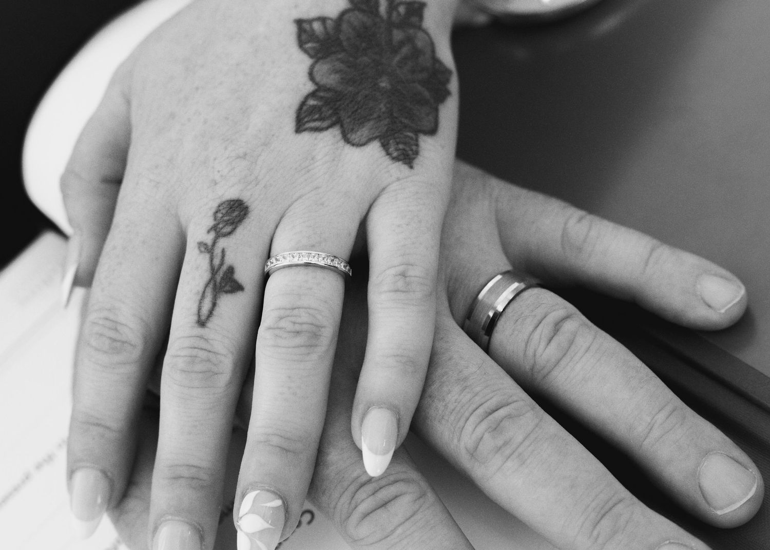 Black and white photo of two hands with wedding rings, one hand featuring floral tattoos, symbolizing marriage.