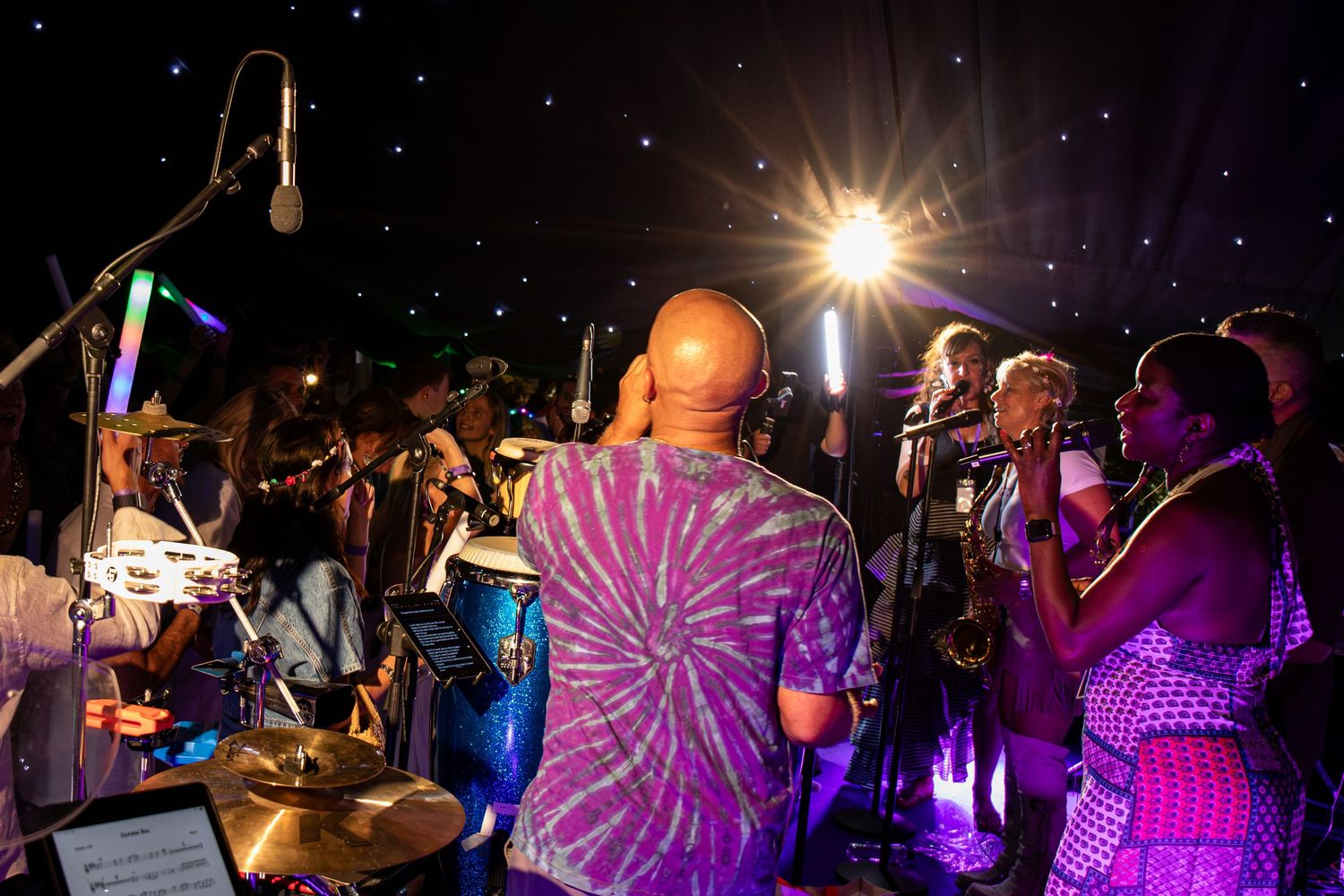 A colorful band performs on stage under starry lights, with a bald man in a tie-dye shirt playing percussion.