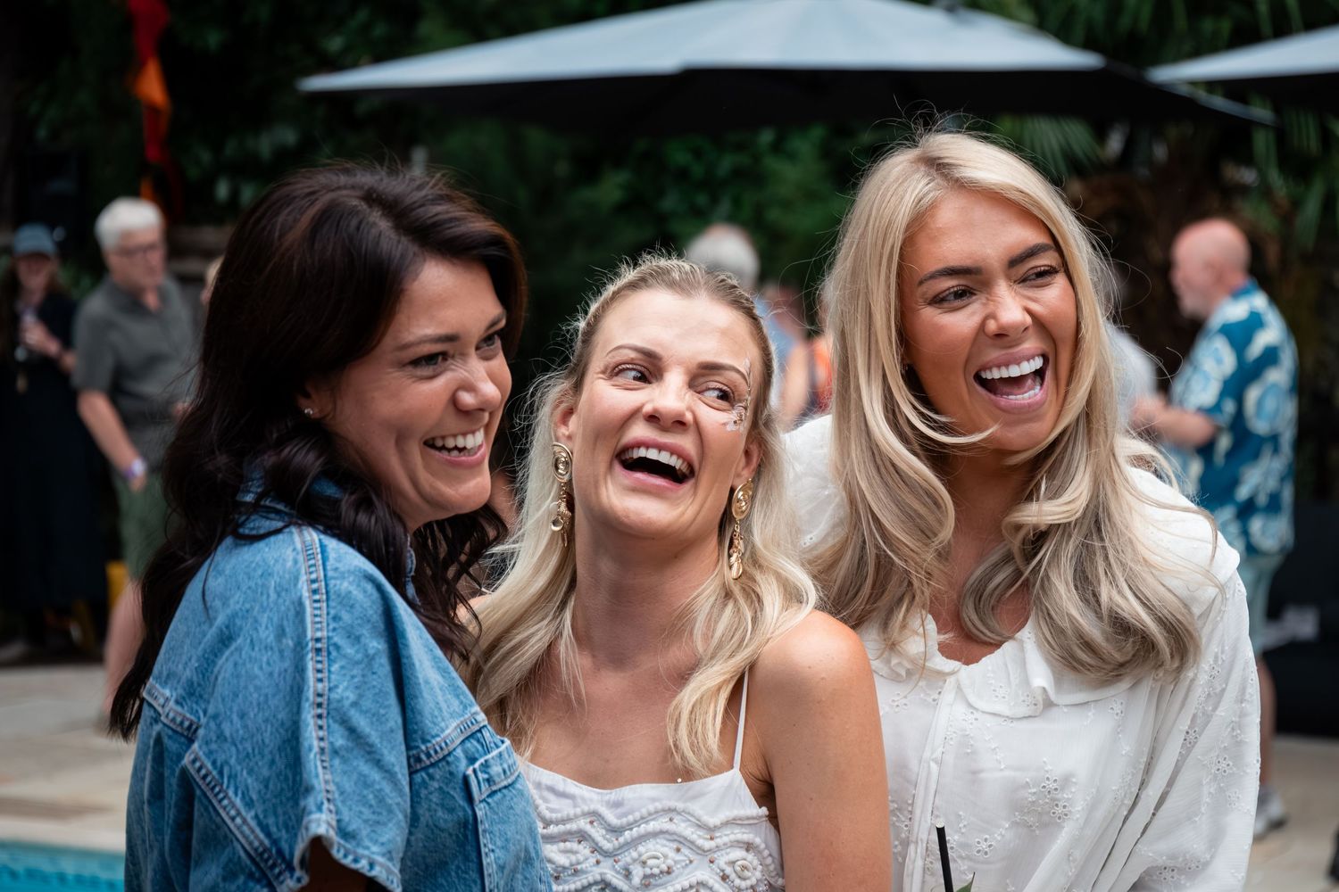 Three happy women laugh together at an outdoor event, two with blonde hair and one with dark hair.