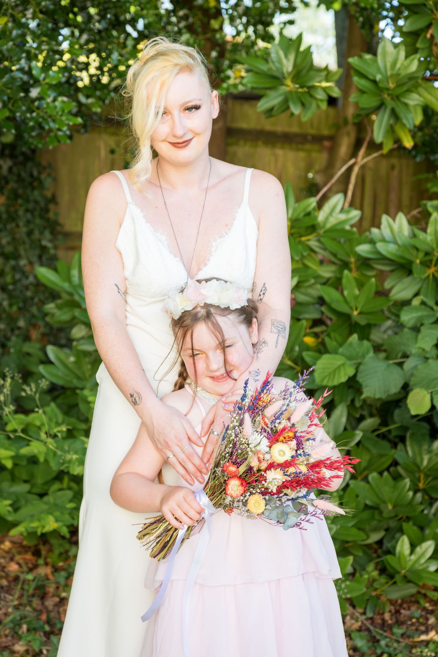 Bride in white dress hugging young flower girl holding colorful bouquet in garden.