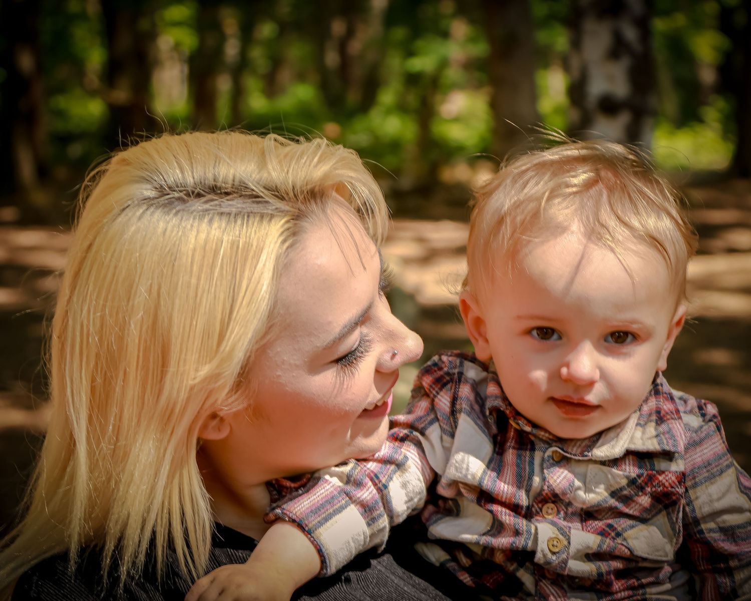Blonde mother gazing lovingly at her toddler son in a plaid shirt in a forest setting.