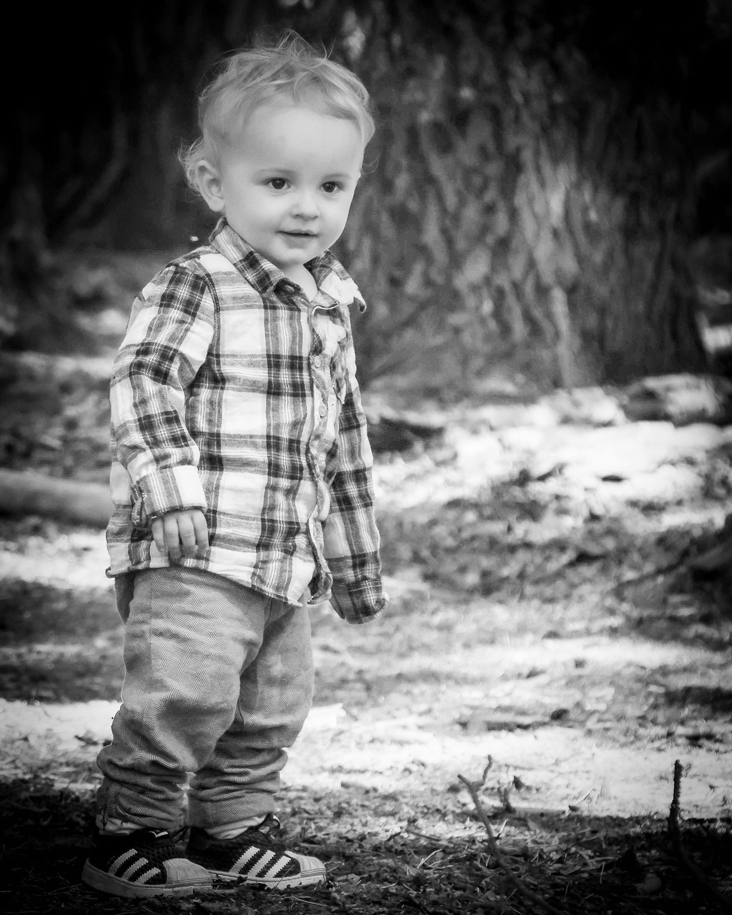 Black and white portrait of a blonde toddler boy in plaid shirt and Adidas shoes outdoors.