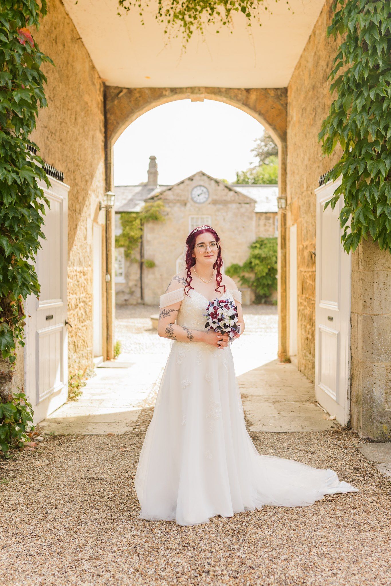Bride in white gown holding bouquet stands under stone arch at elegant venue.