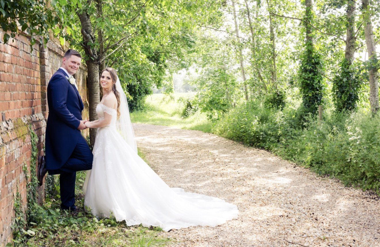 Bride and groom posing on a tree-lined gravel path, bride in flowing white gown with long veil.