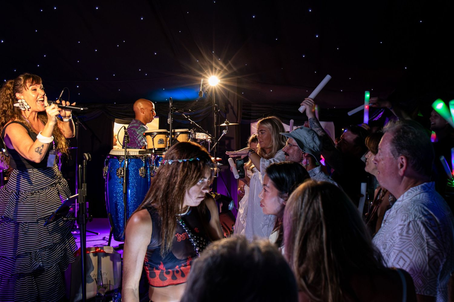 A lively band performs on a colorful stage as an energetic crowd celebrates at an indoor night event.