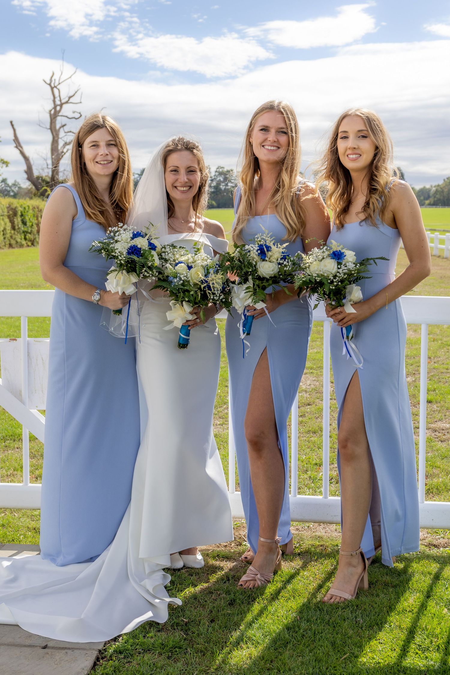 Bride in white gown with three bridesmaids in light blue dresses holding white and blue bouquets outdoors.