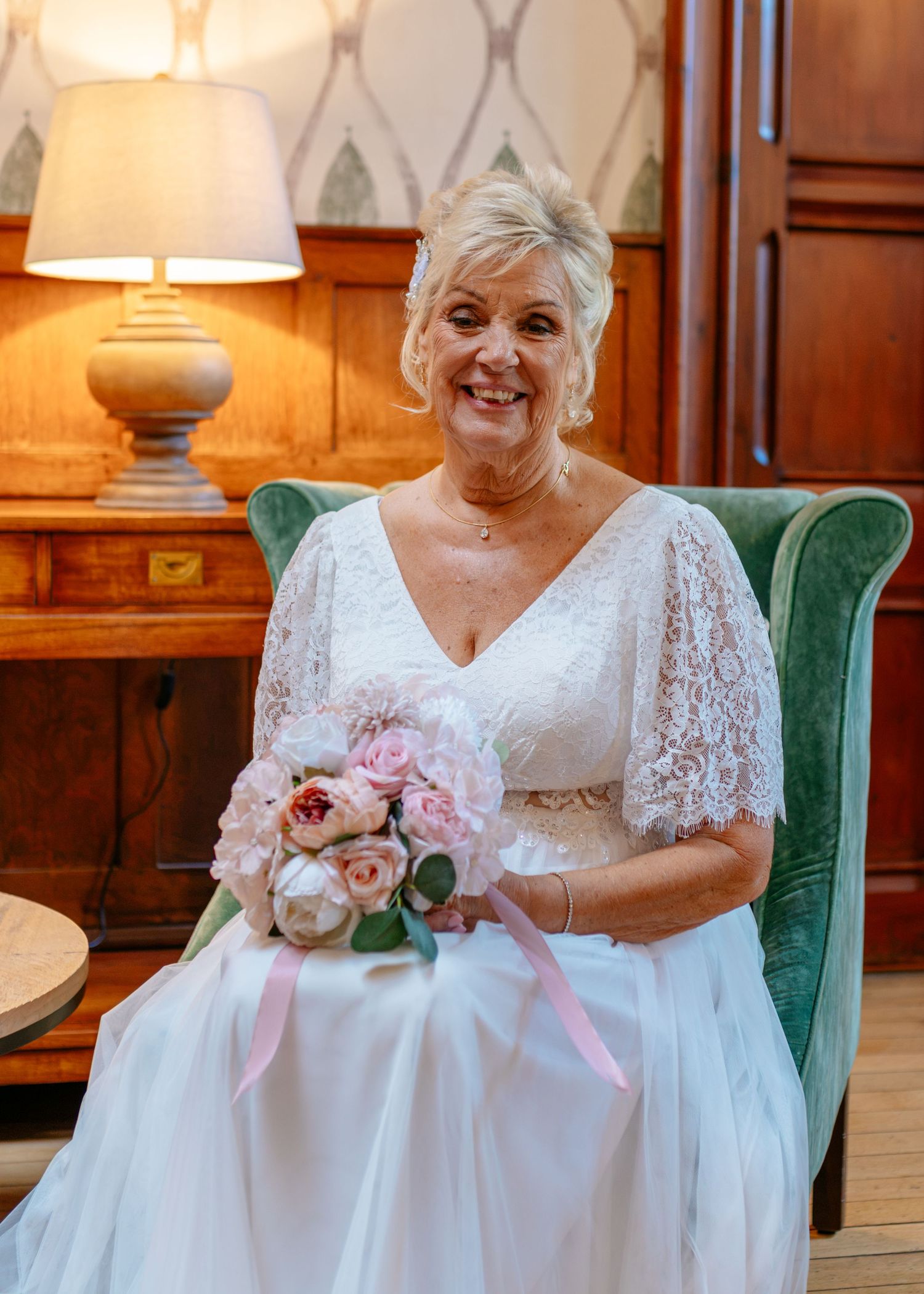 Smiling bride in white lace dress holding pink bouquet seated in green velvet chair.