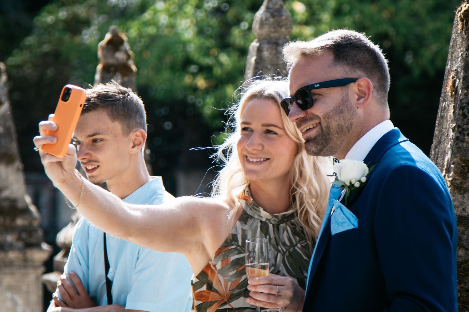 Three people taking a selfie at a wedding, with the groom in a blue suit and boutonniere smiling outdoors.