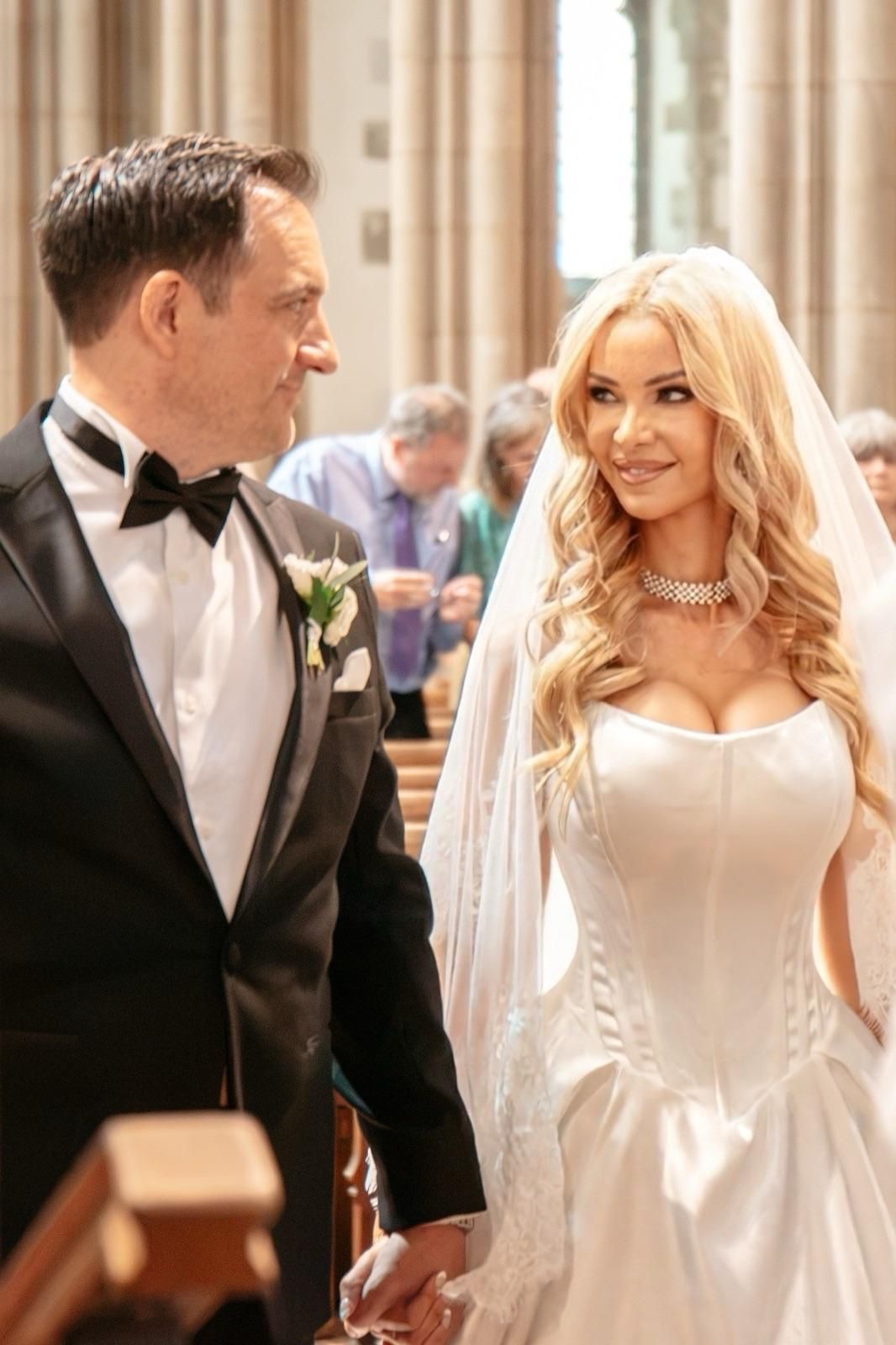 Elegant bride in white corset gown and veil holds hands with groom in black tuxedo at church wedding ceremony.