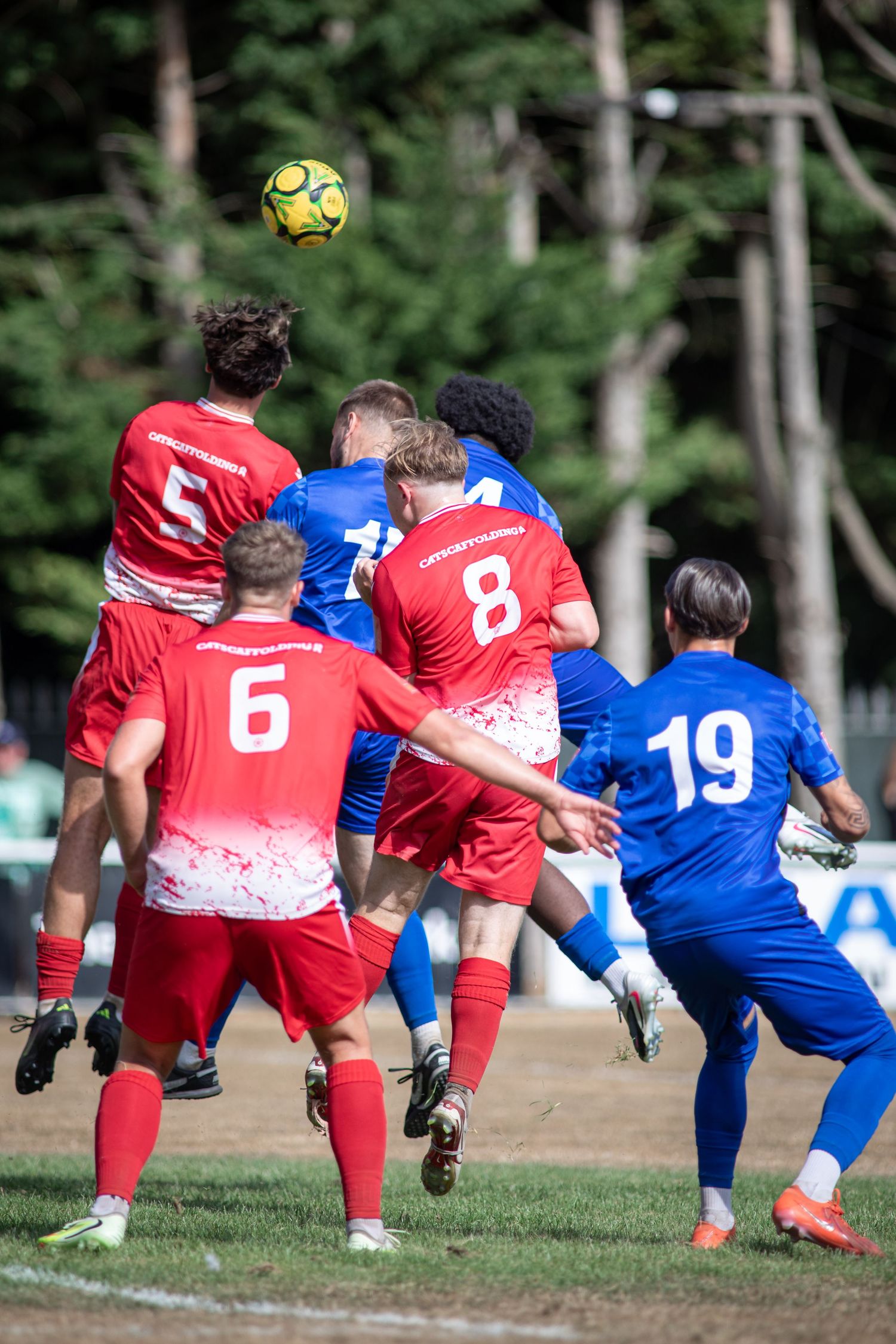 Red and blue soccer players contest aerial header during sunny outdoor football match.
