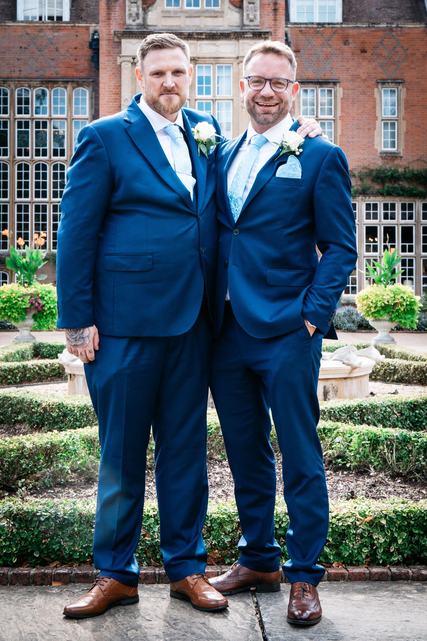 Two grooms in matching navy blue suits with boutonnieres pose together outside a grand manor house venue.