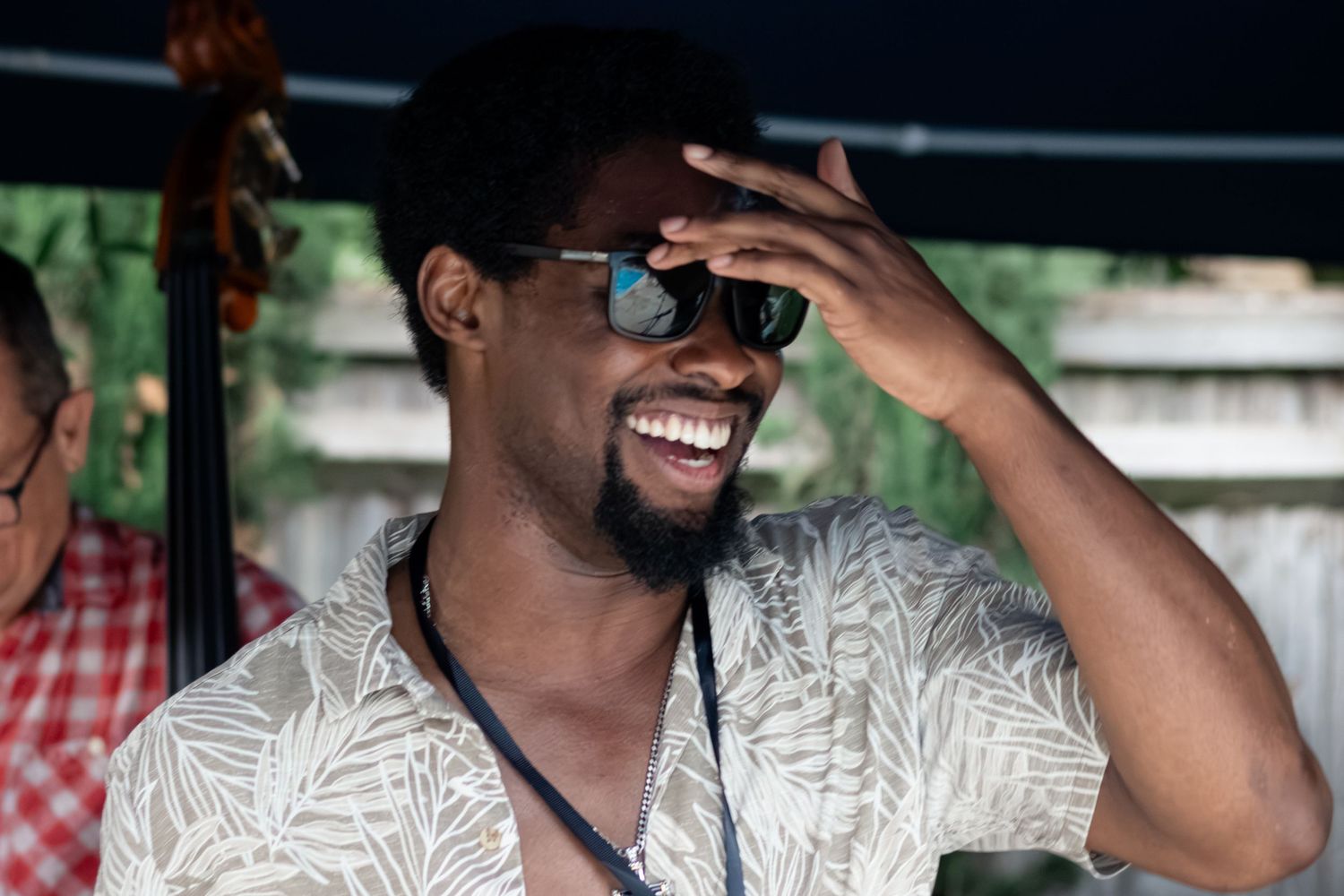 A smiling Black man wearing sunglasses and a floral shirt laughs joyfully at an outdoor music event.