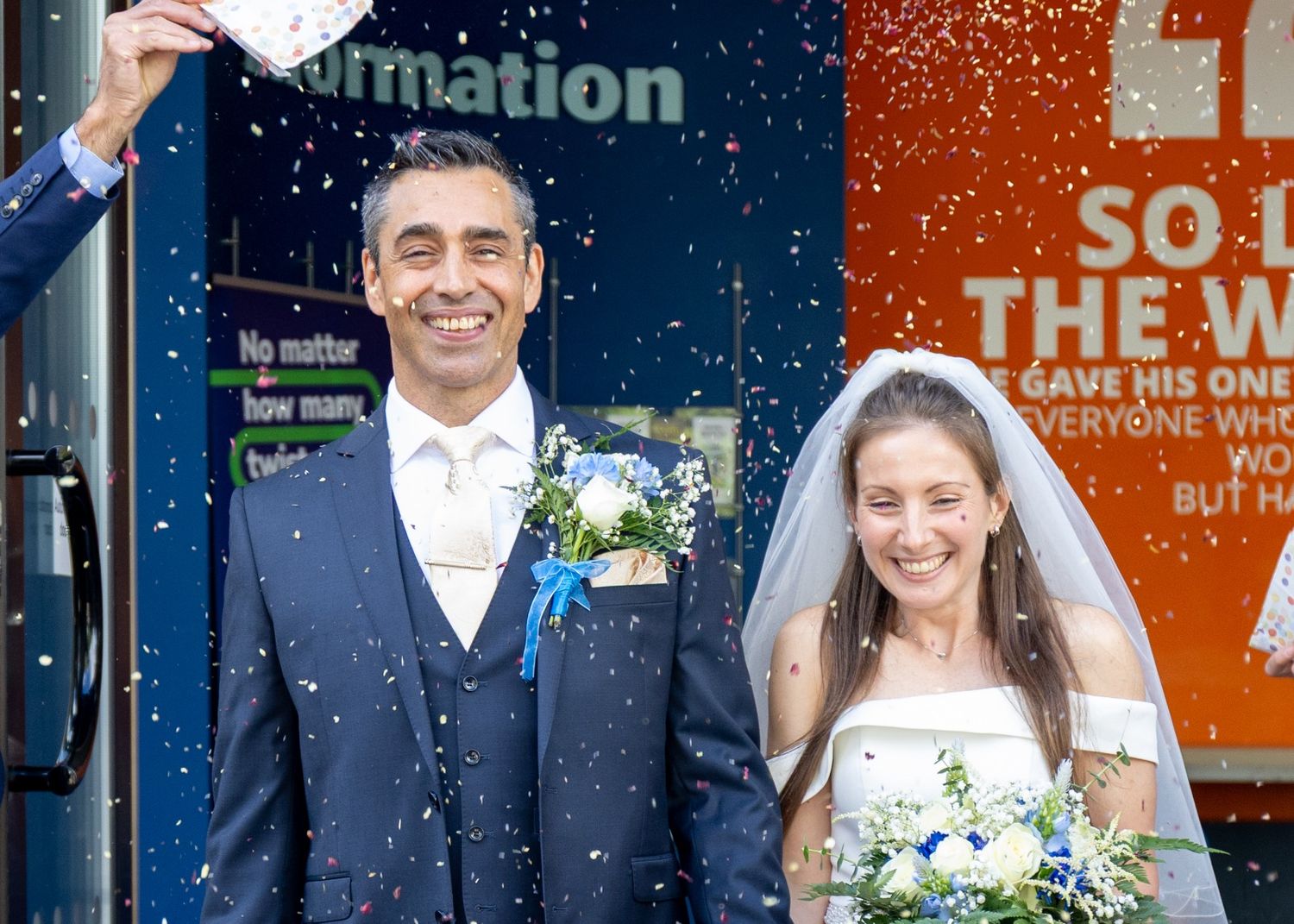 Happy couple celebrates wedding outdoors with confetti, groom in navy suit, bride in white gown holding bouquet.