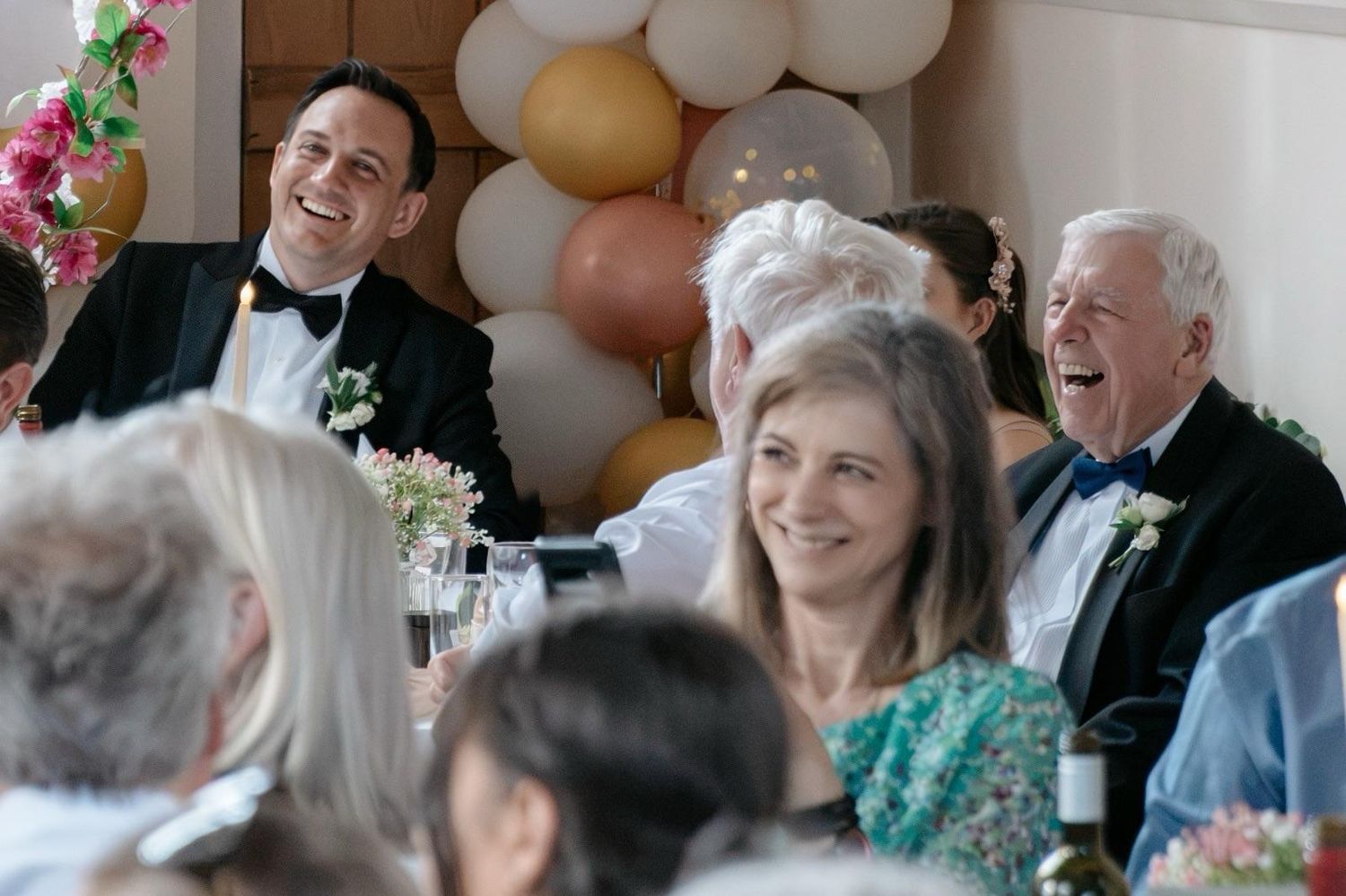 Wedding guests laugh and smile during joyful reception dinner with floral decorations and balloons.