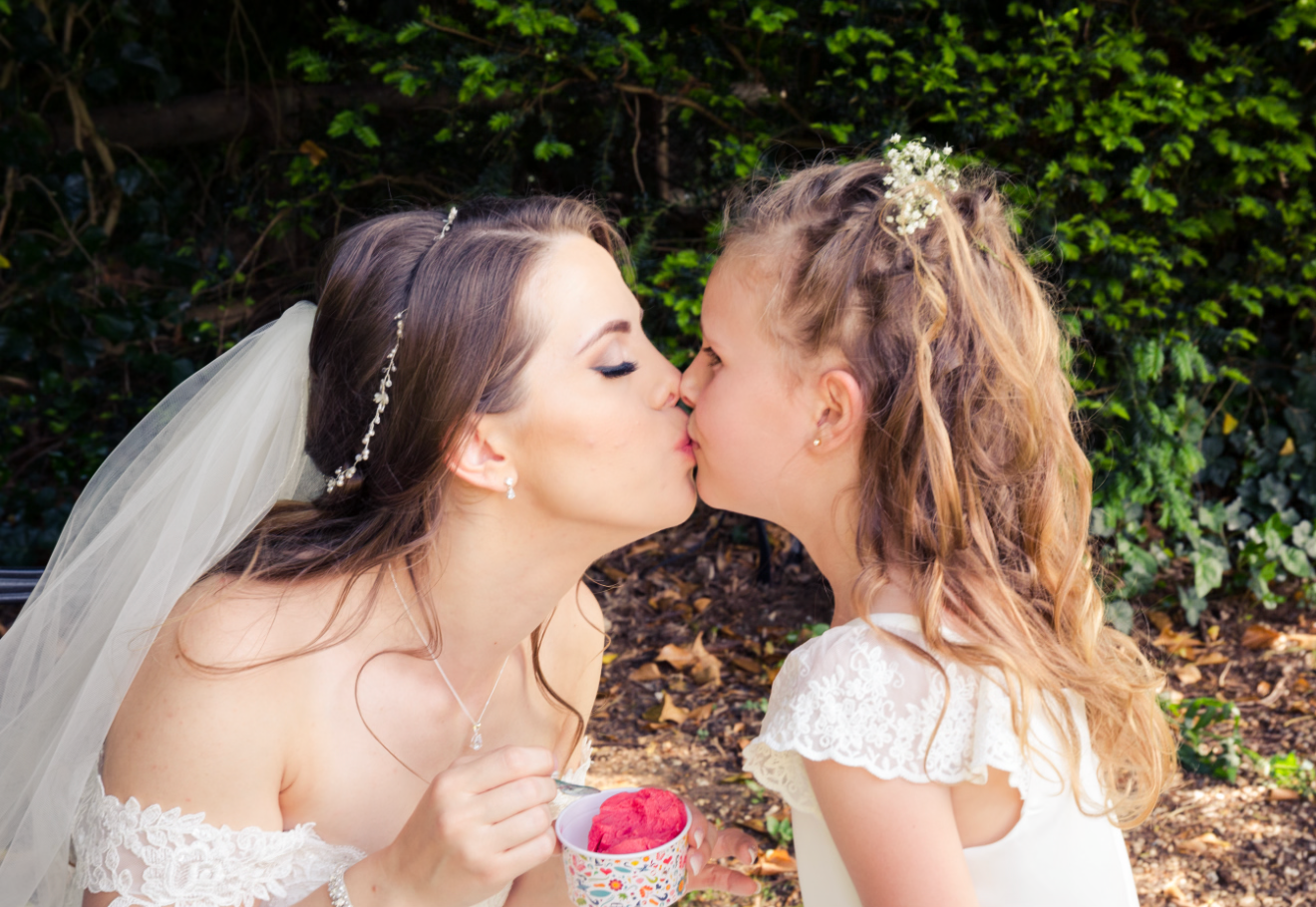 Bride sharing a tender kiss with flower girl in white dress at outdoor wedding ceremony.