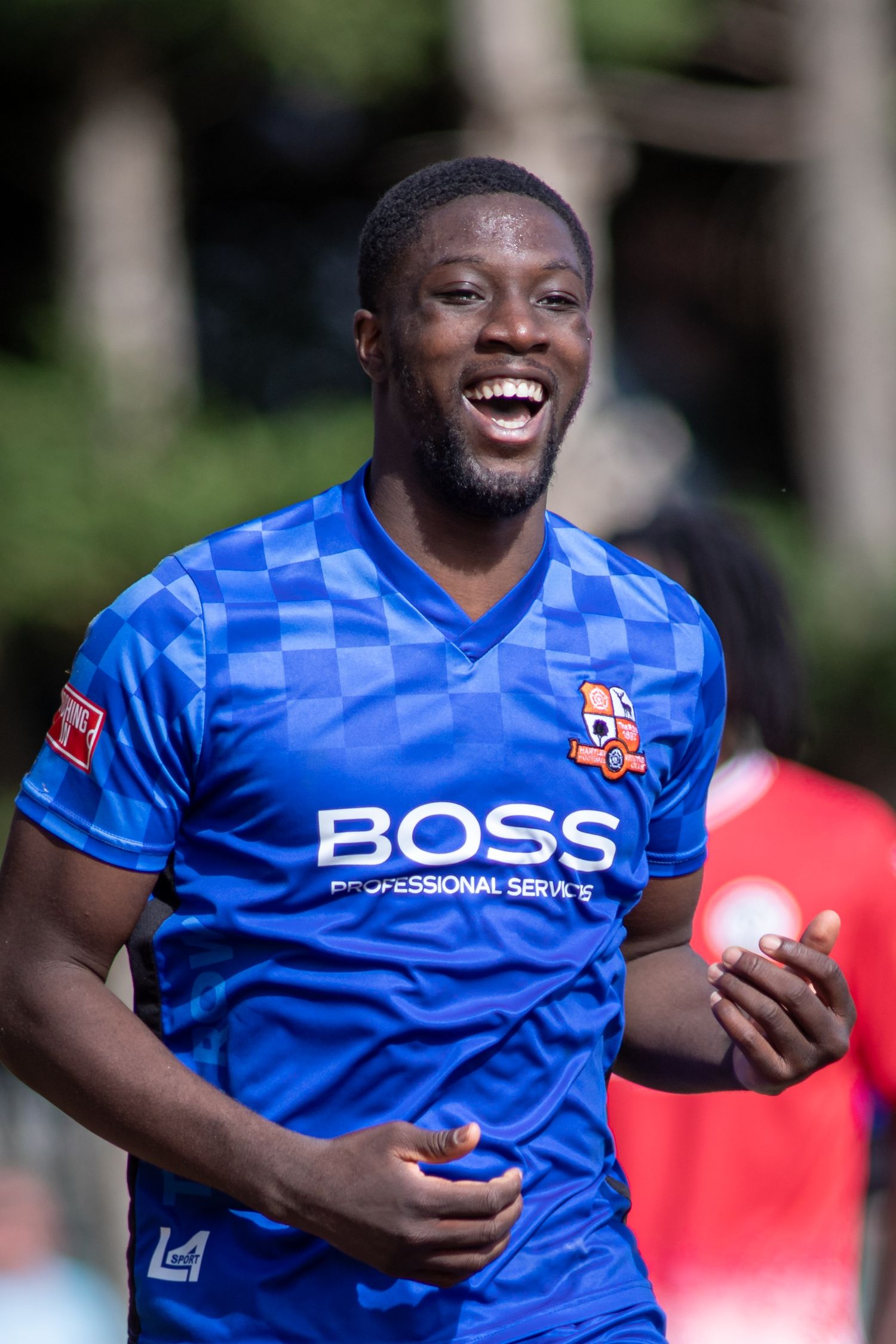 Smiling footballer in blue BOSS sponsored checkered kit celebrates during a sunny outdoor match.