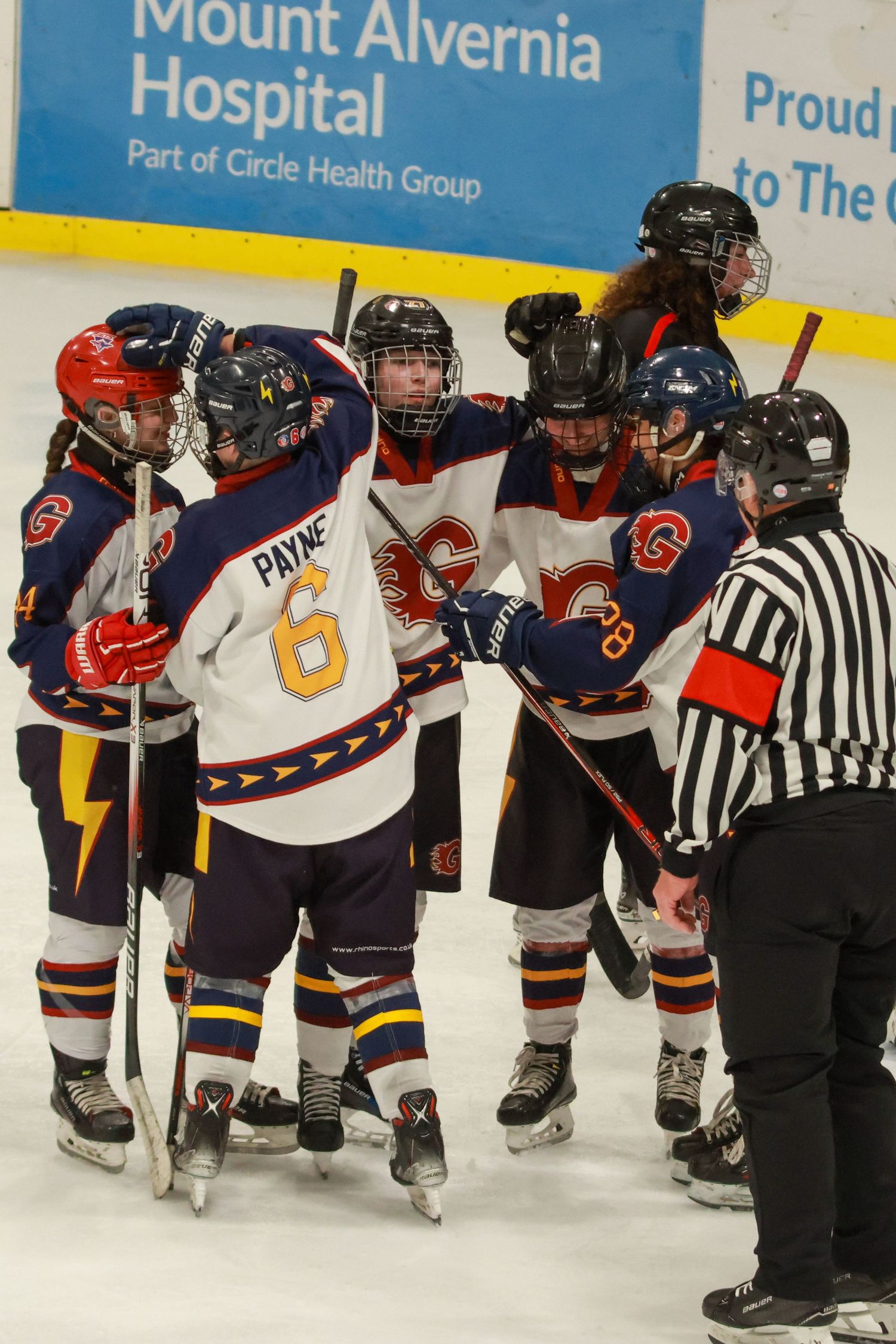 Female ice hockey team celebrates goal on ice, player Payne number 6 visible, referee nearby, Mount Alvernia Hospital banner behind.