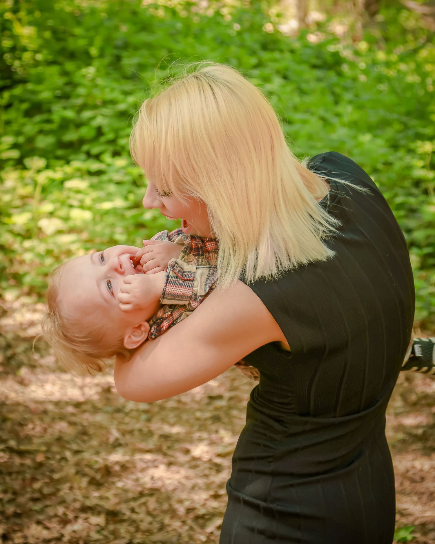 Joyful blonde mother lifting and playing with her laughing toddler boy outdoors in a forest.