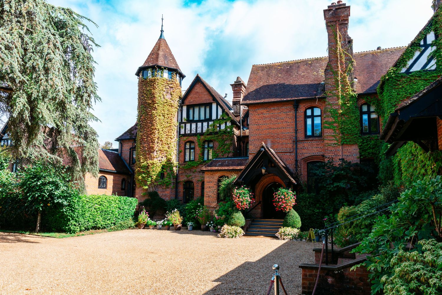 Charming red brick Tudor manor house with ivy-covered walls, round tower, and manicured topiary in a scenic English countryside setting.