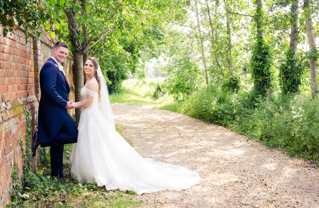 Bride and groom holding hands on a tree-lined path, bride in white gown with veil, groom in navy suit.