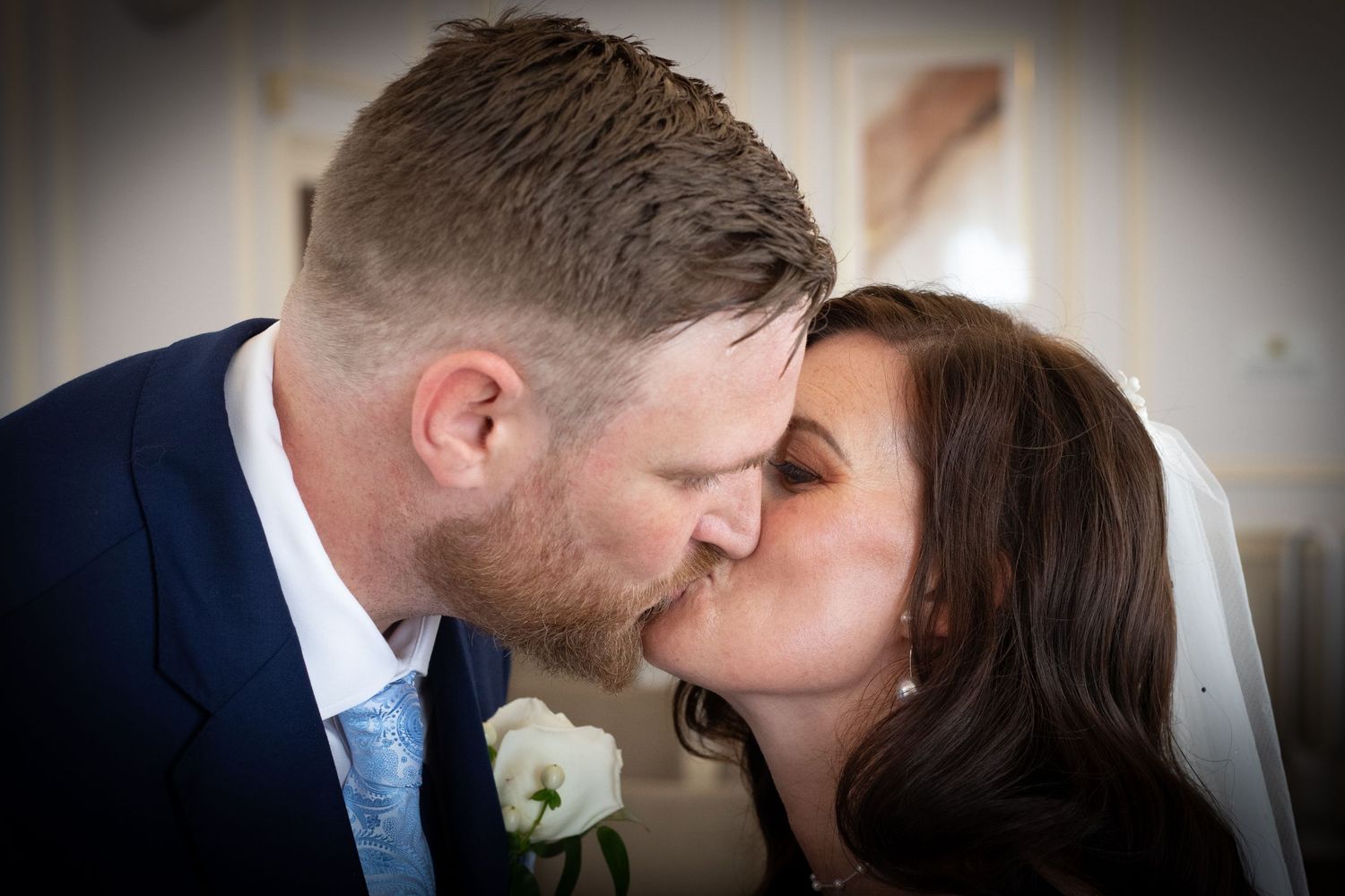 Bride and groom share a romantic kiss on their wedding day, with the groom in a navy suit and white boutonniere.