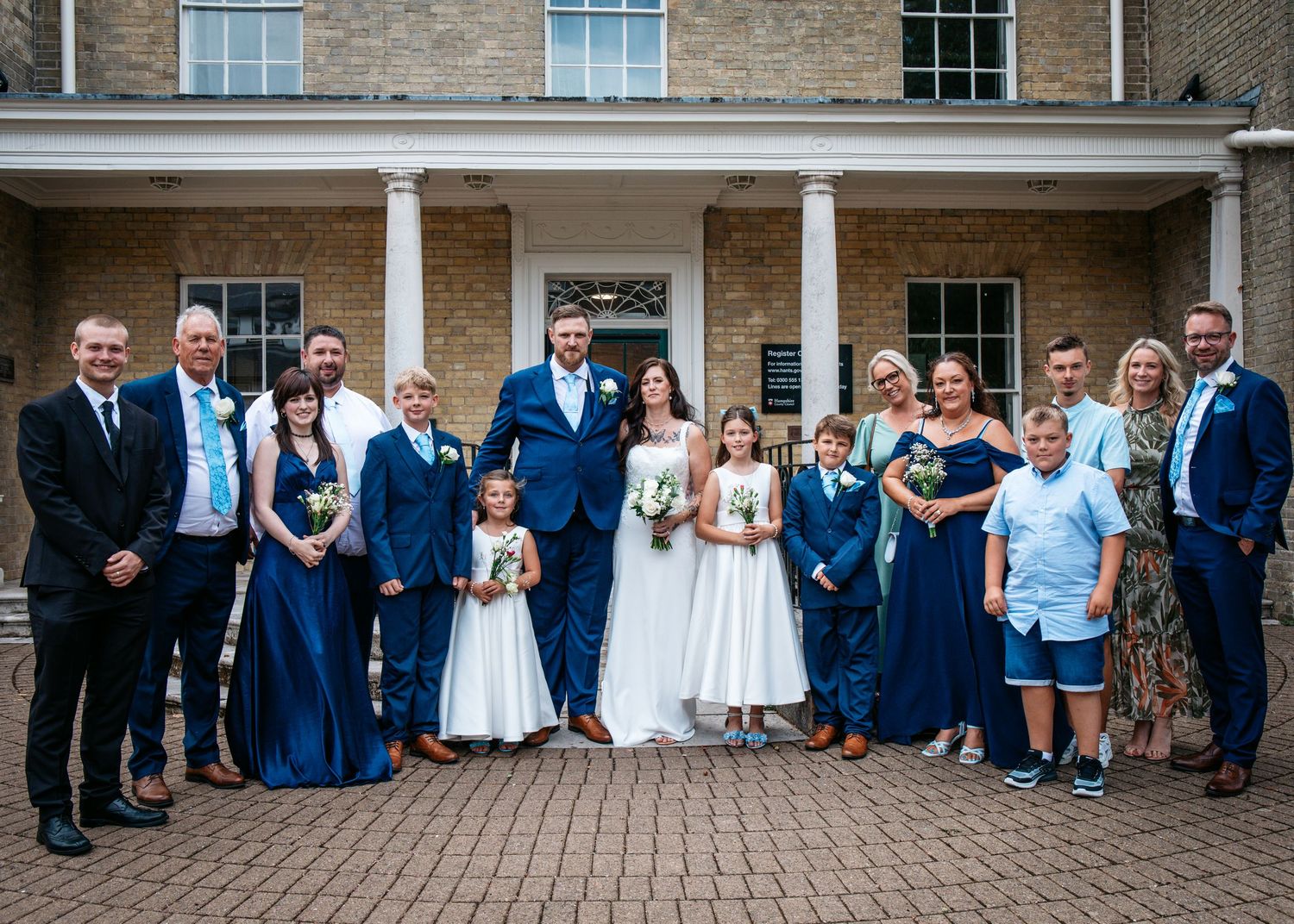 Wedding party poses outside elegant venue, bride in white gown with groom and guests in navy blue formal attire.