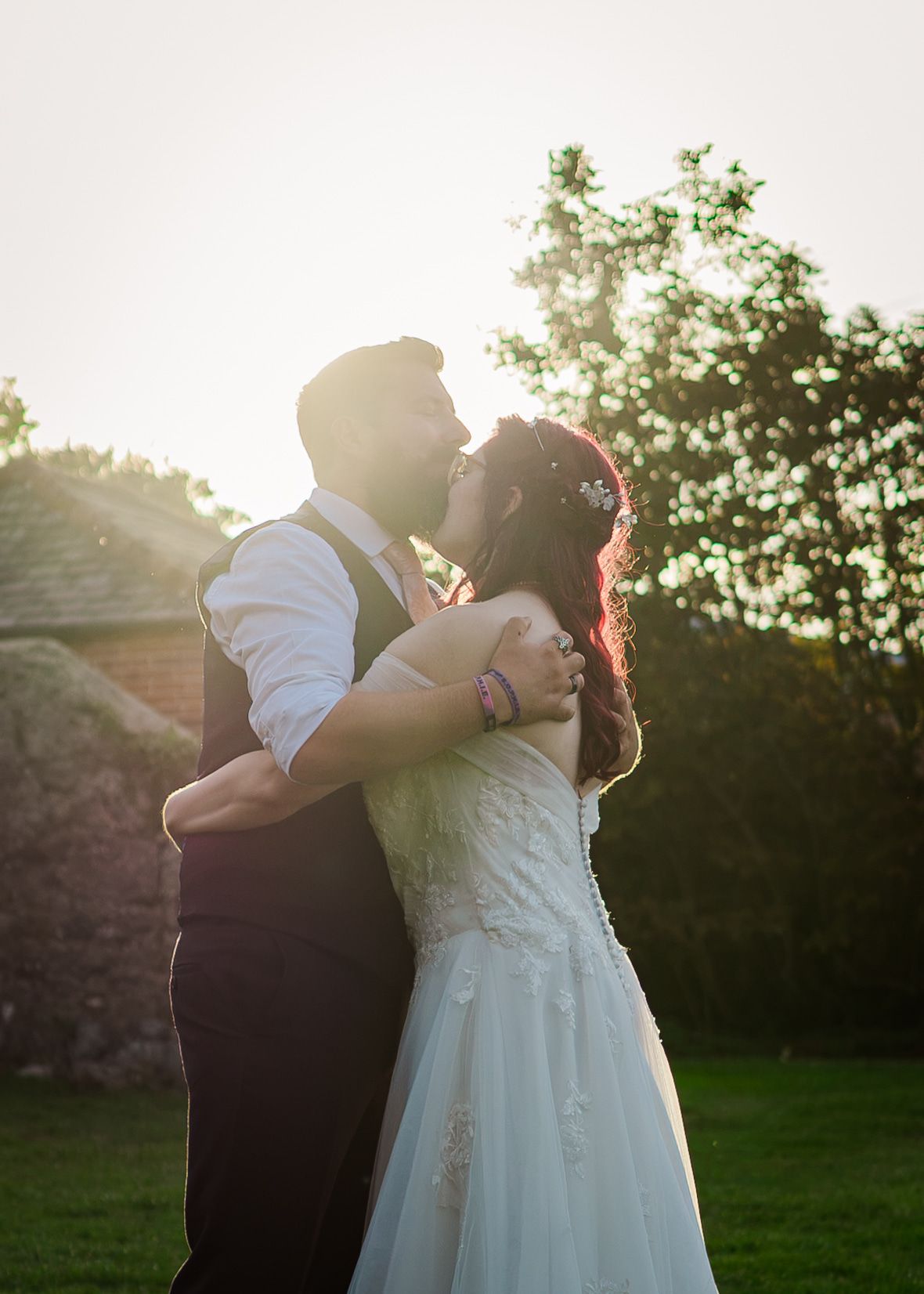 Bride and groom embrace in golden sunset light outdoors, wearing white wedding dress and vest, surrounded by lush green trees.