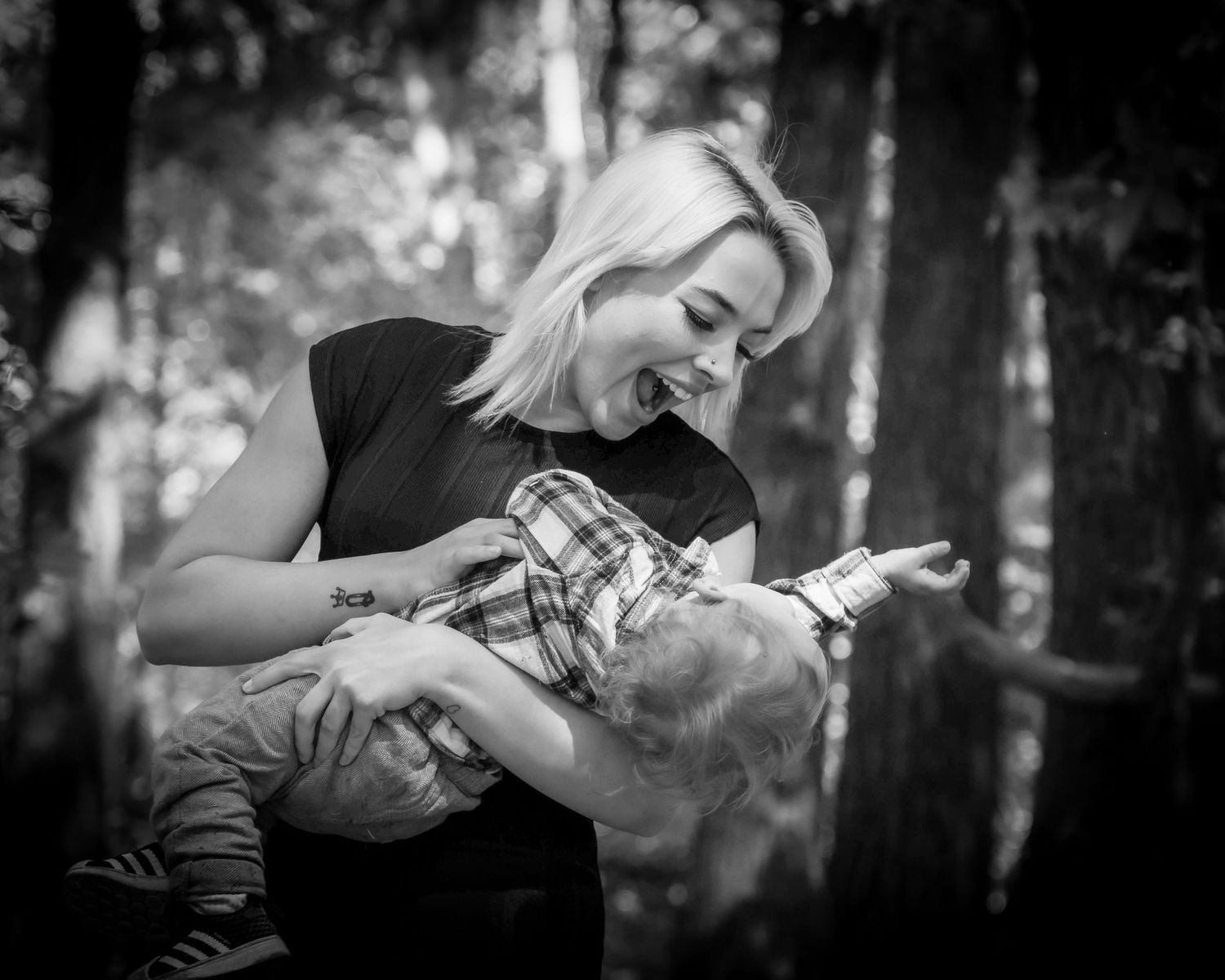 Black and white photo of a laughing mother joyfully lifting her toddler son in a forest.