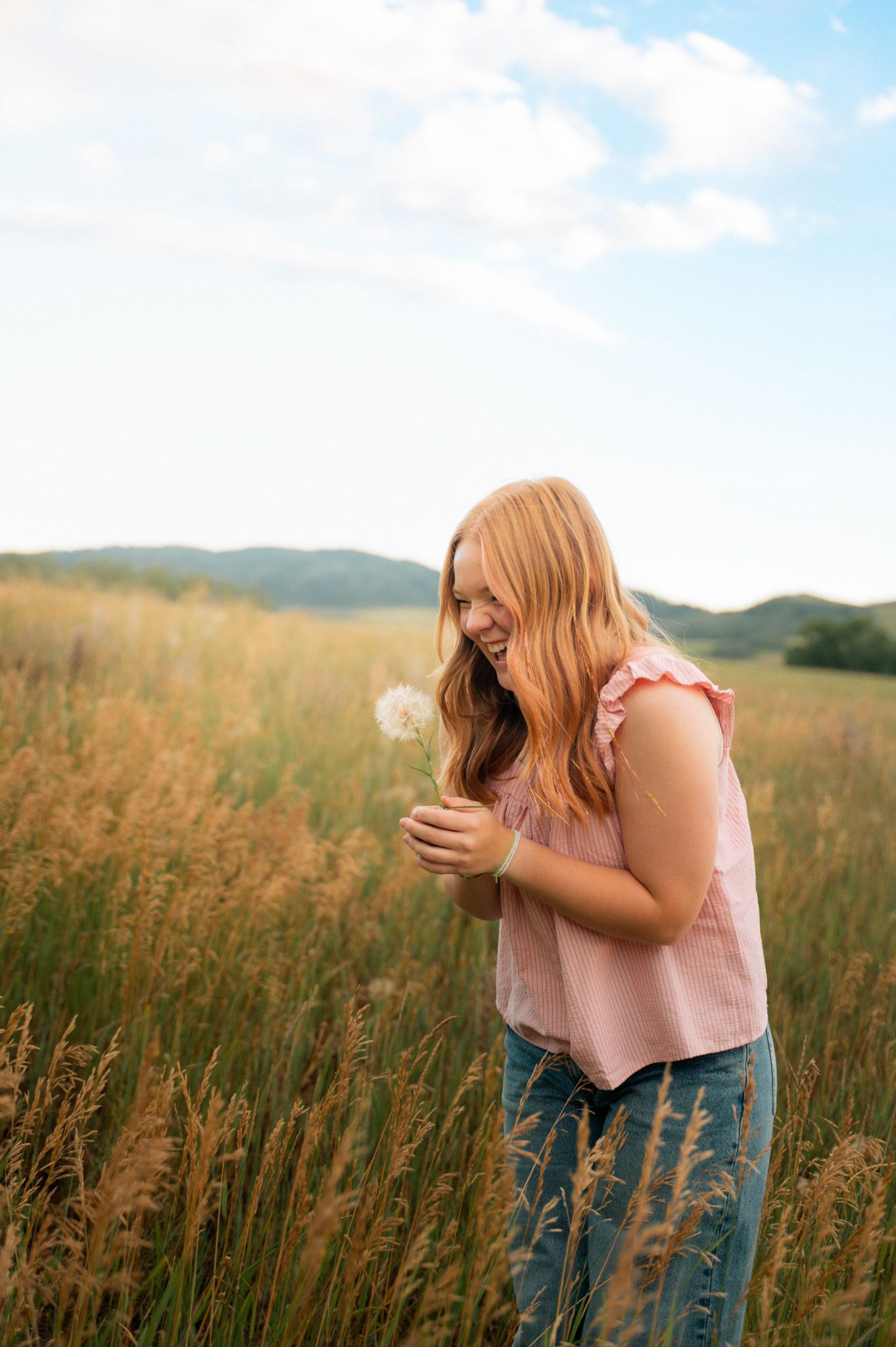 A smiling teenage girl with red hair holds a dandelion in a golden meadow under blue skies.