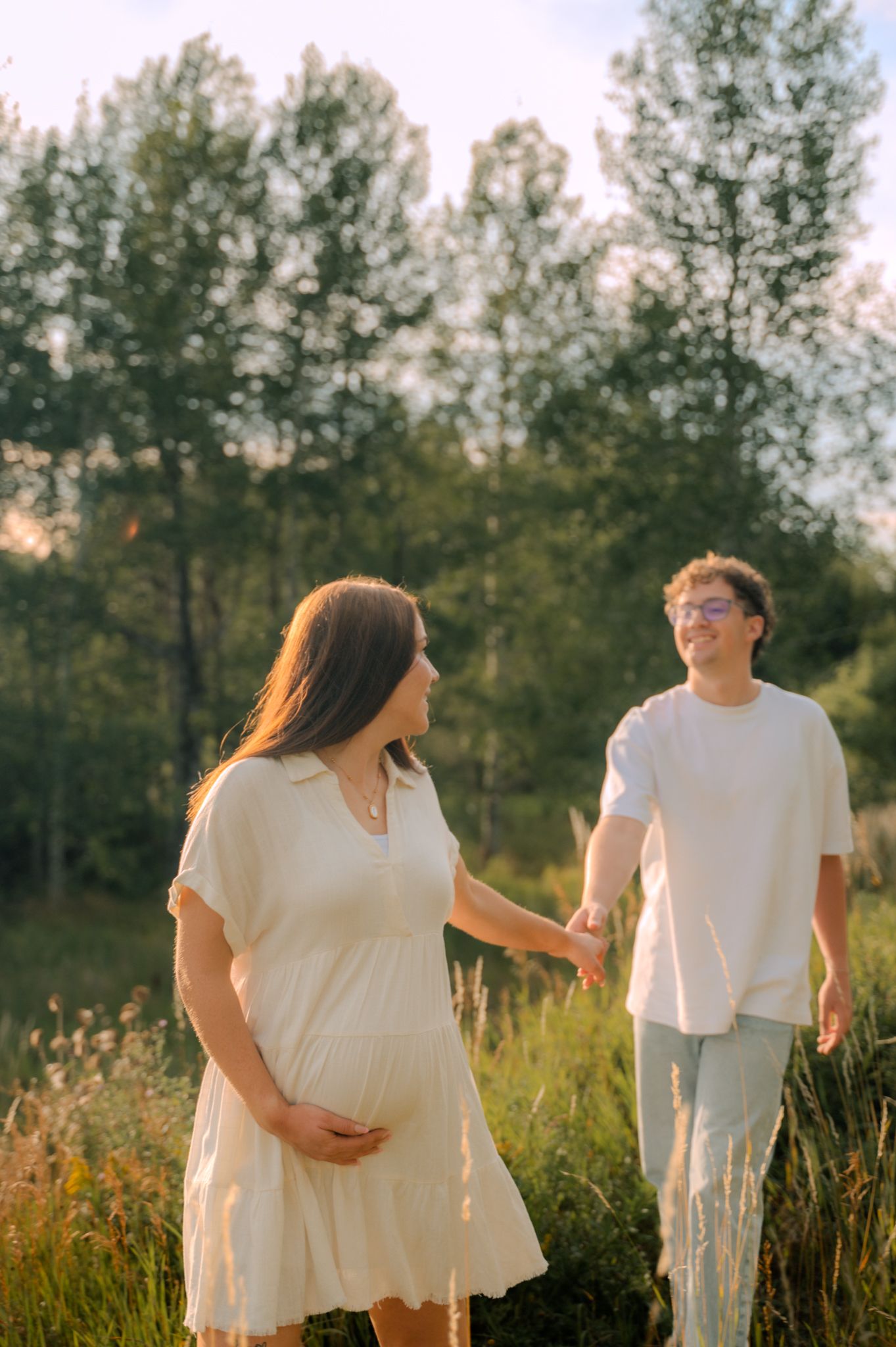 Pregnant woman in white dress holding belly while partner holds her hand in sunny green field.