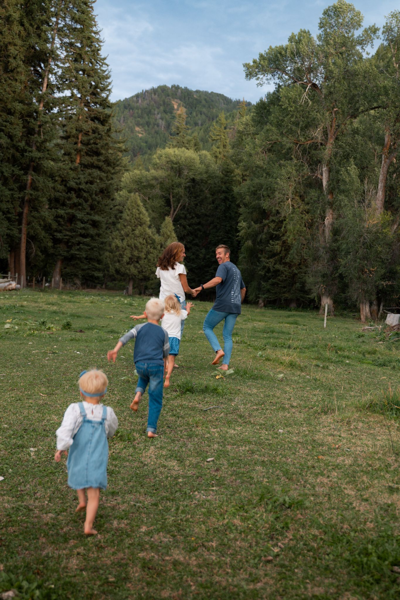 Family with three young children running through a lush green meadow surrounded by tall pine trees and mountains.