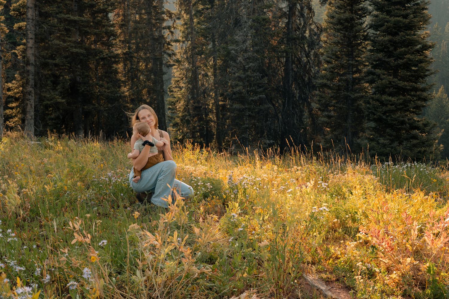 A woman sits smiling in a golden sunlit alpine meadow with tall evergreen trees behind her.