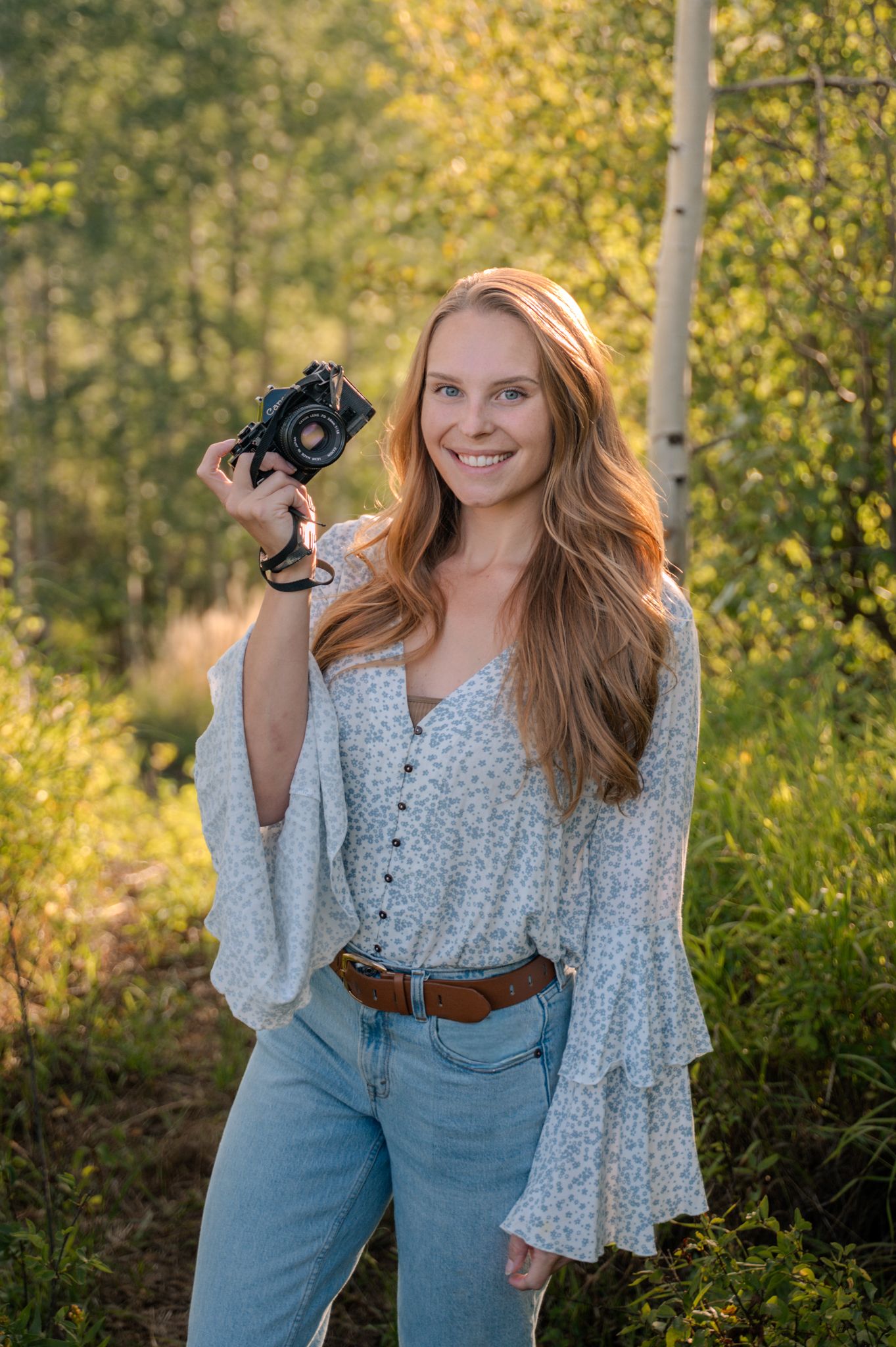 Smiling young woman with long auburn hair holding camera in sunlit aspen forest setting.