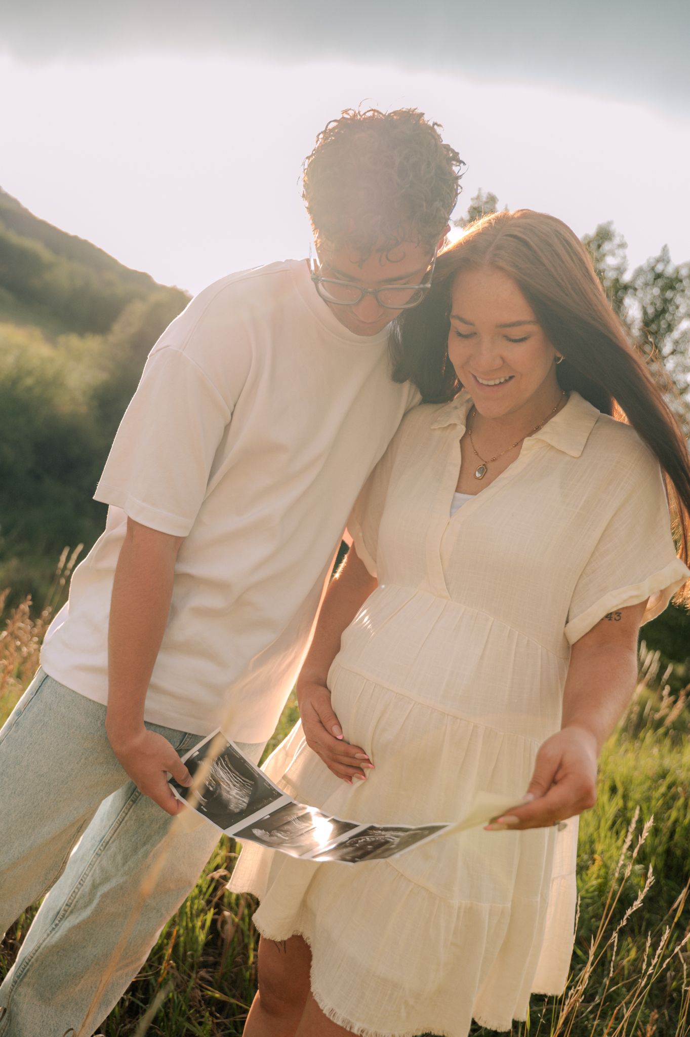 Happy couple reviewing ultrasound photos outdoors in a sunny green field at golden hour.