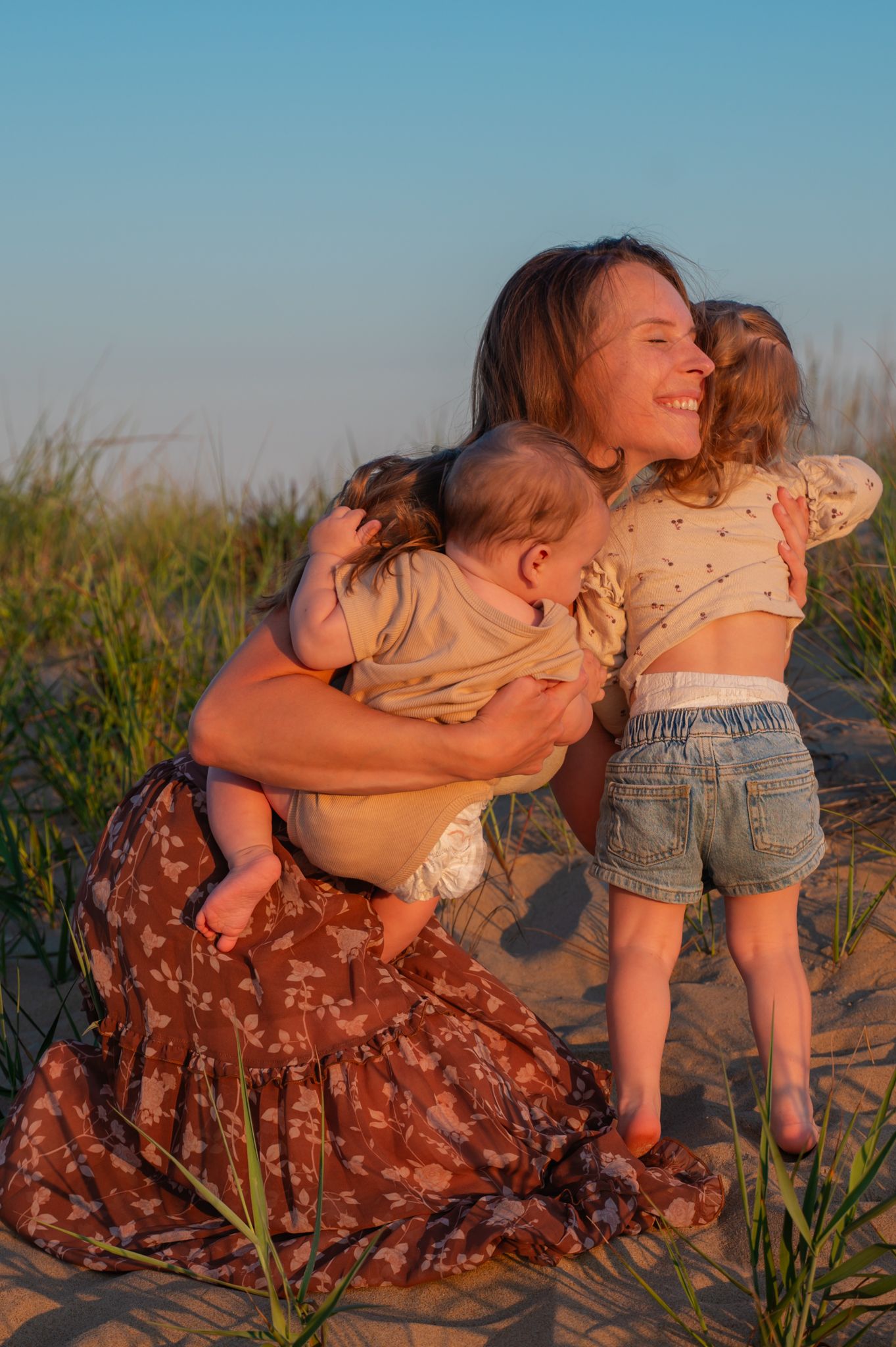 A smiling mother holds her baby while her toddler hugs her on a sandy beach at golden hour.