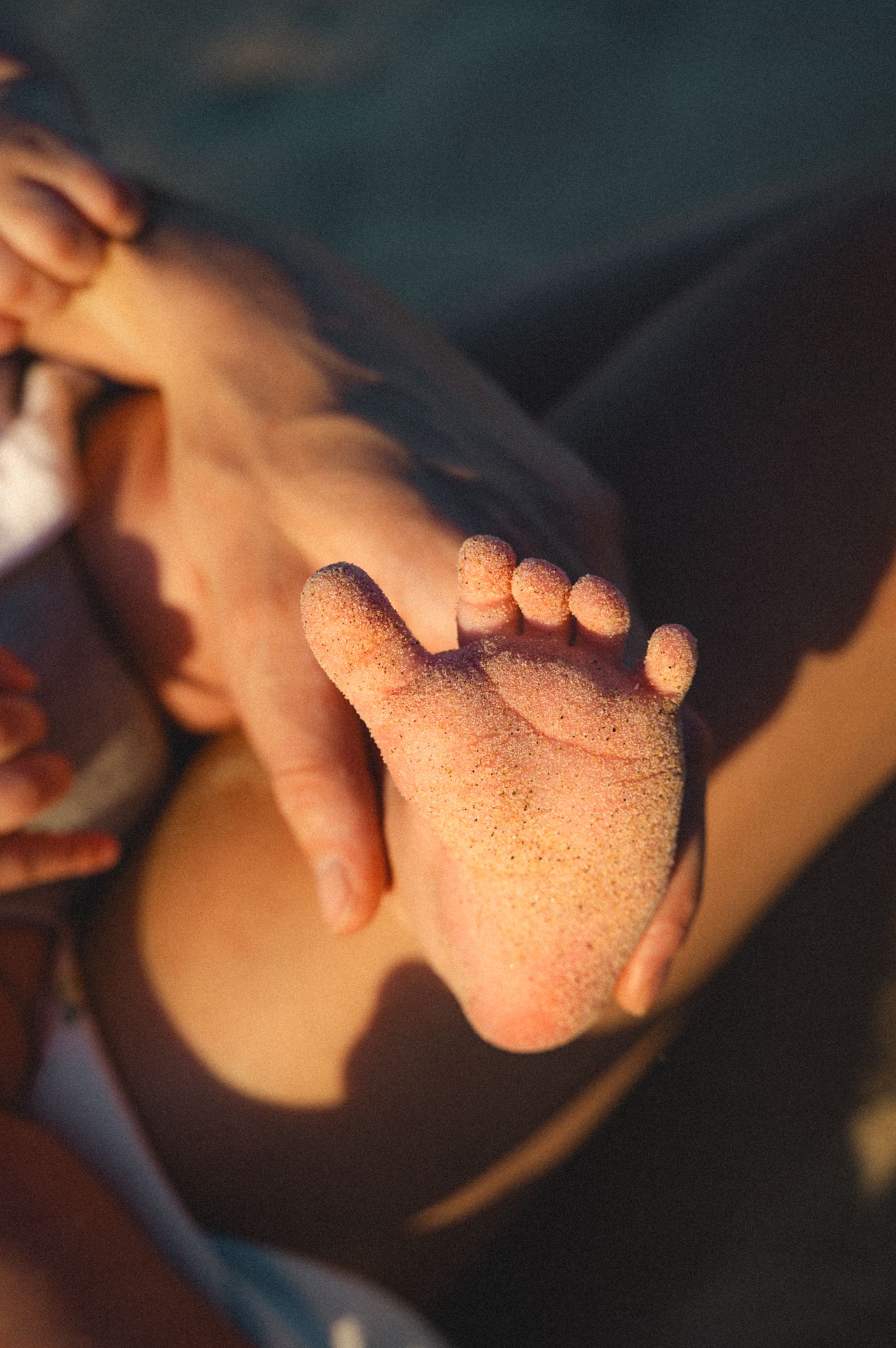 Close-up of a sandy baby foot held gently by a parent's hand at the beach.