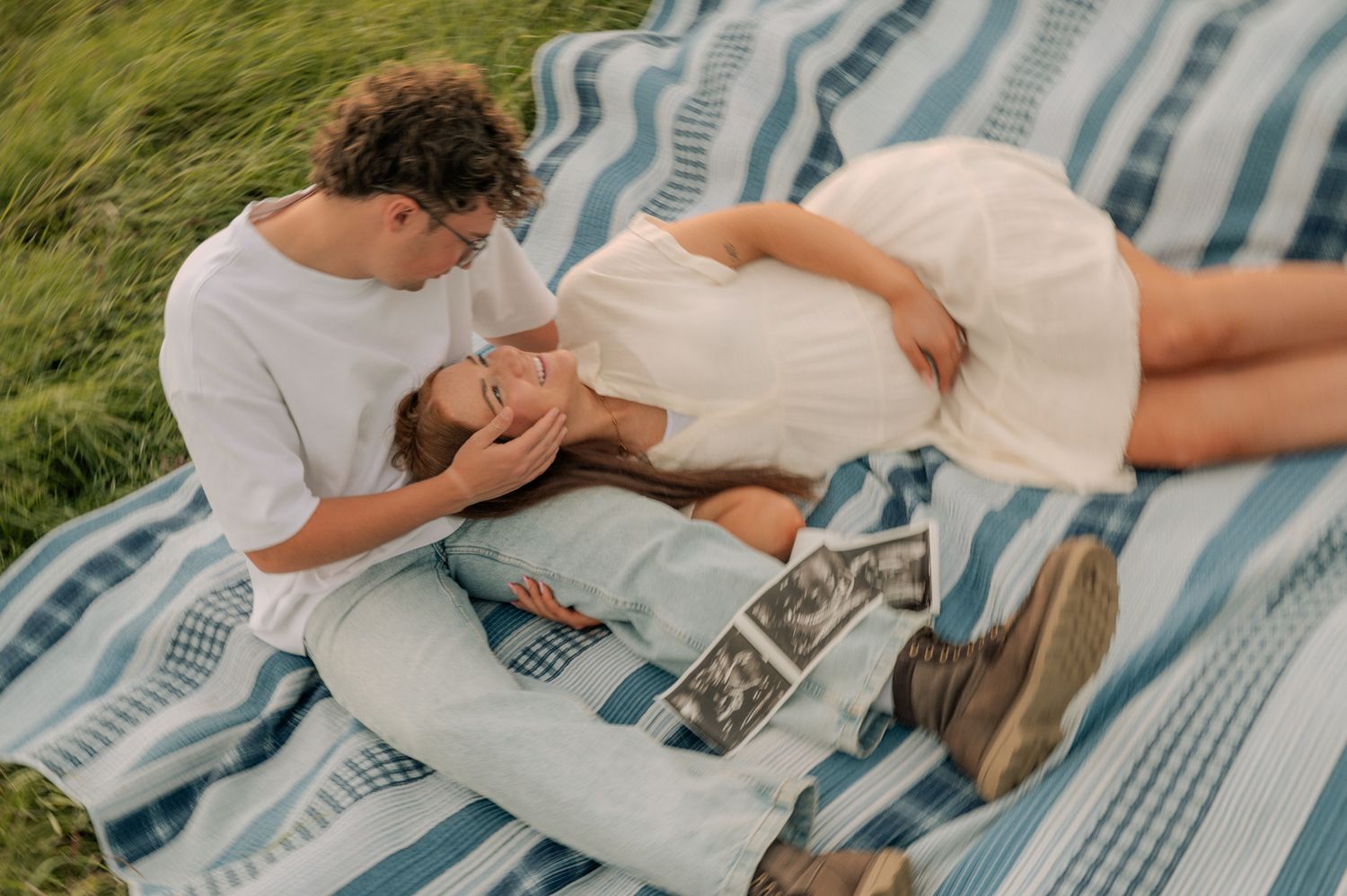 Couple relaxing on striped blanket in grass with ultrasound photos, woman lying on mans lap.