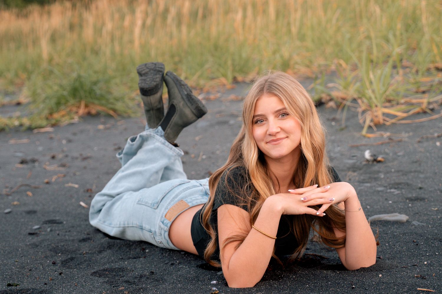 Young blonde woman in jeans and black top lying on dark sand beach, smiling at camera.
