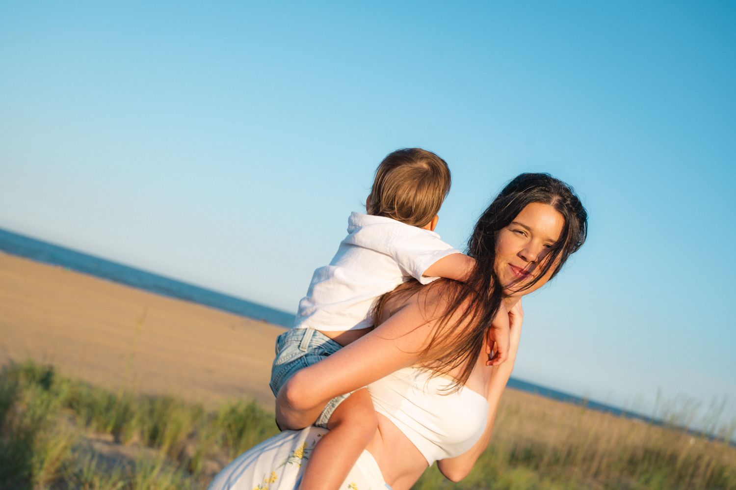 Mother carrying young child on her back in a sunny coastal grass field.