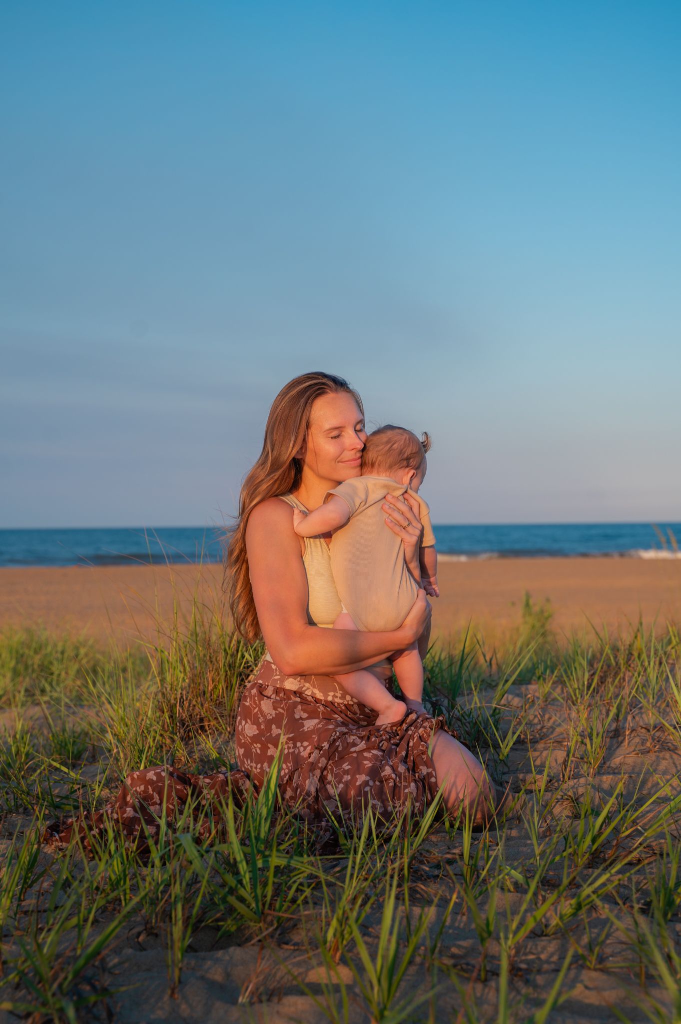 Mother sitting on beach dunes at sunset cradling and kissing newborn baby with ocean backdrop.