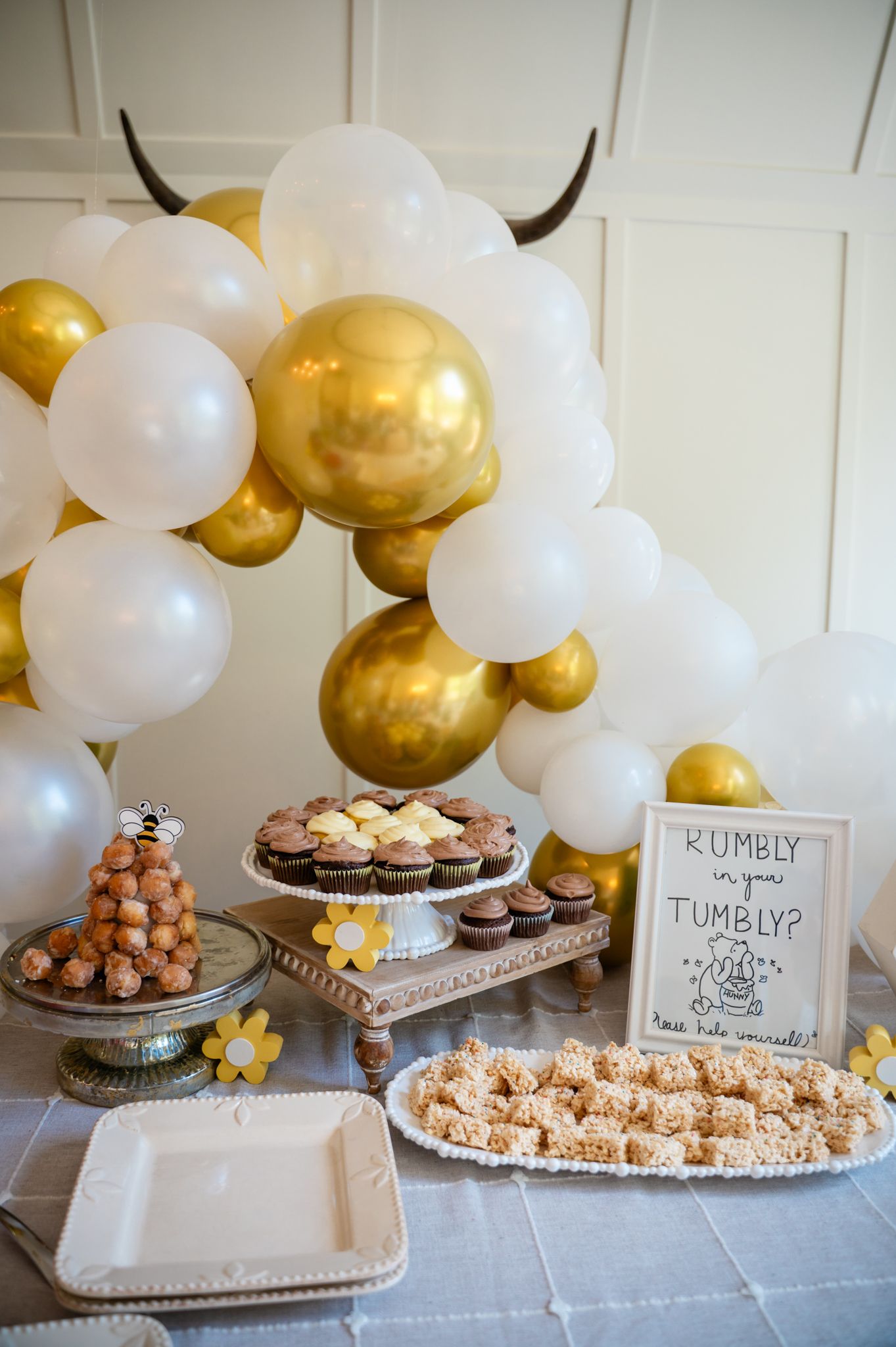 Gold and white balloon arch over dessert table with cupcakes and treats at baby shower.