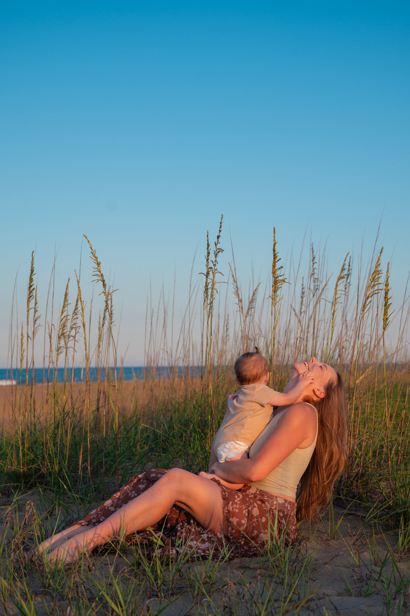 Mother sitting in beach grass at sunset holding baby while leaning back joyfully.