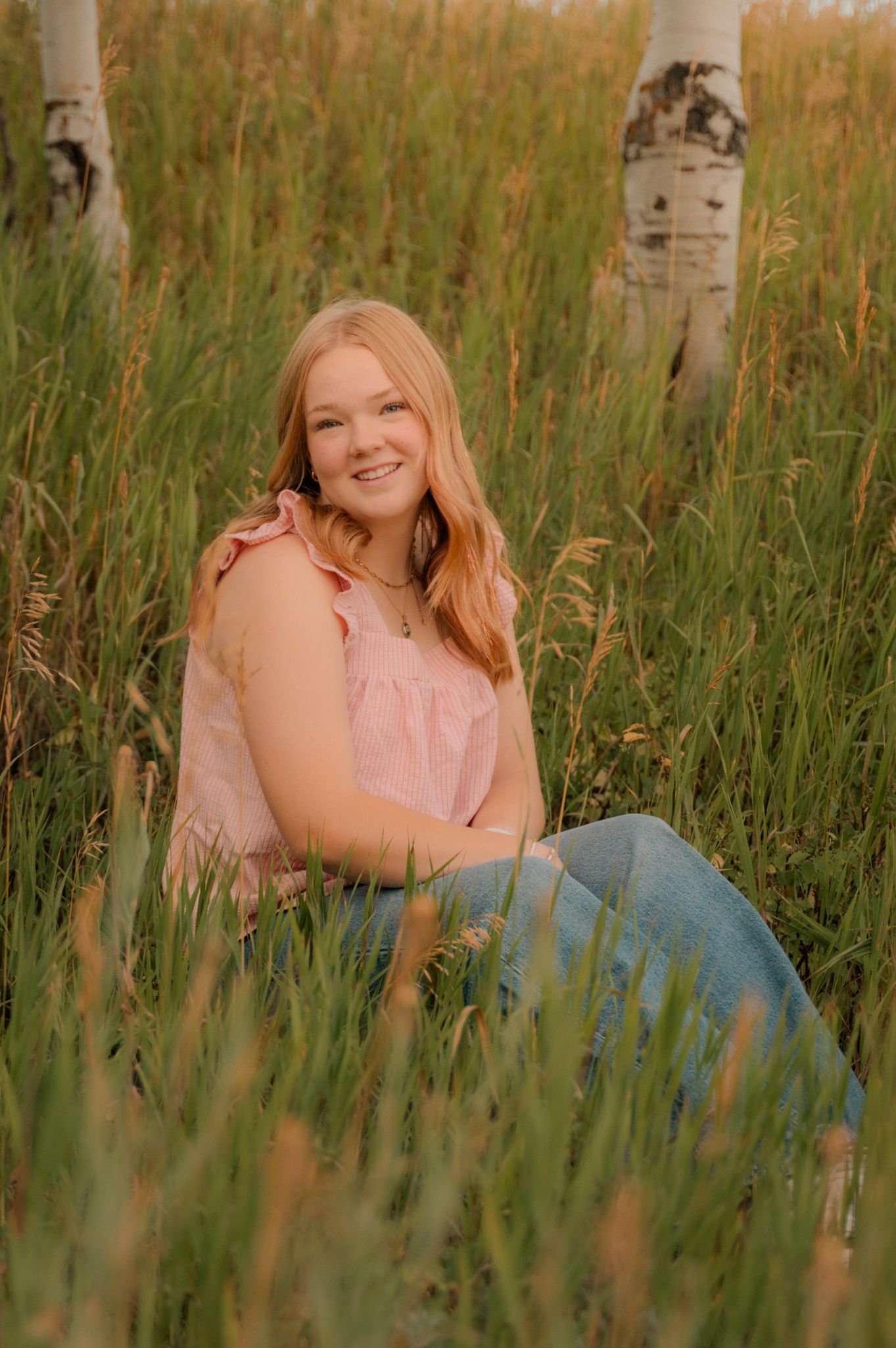 Smiling teenage girl sitting in tall grass near birch trees at sunset.