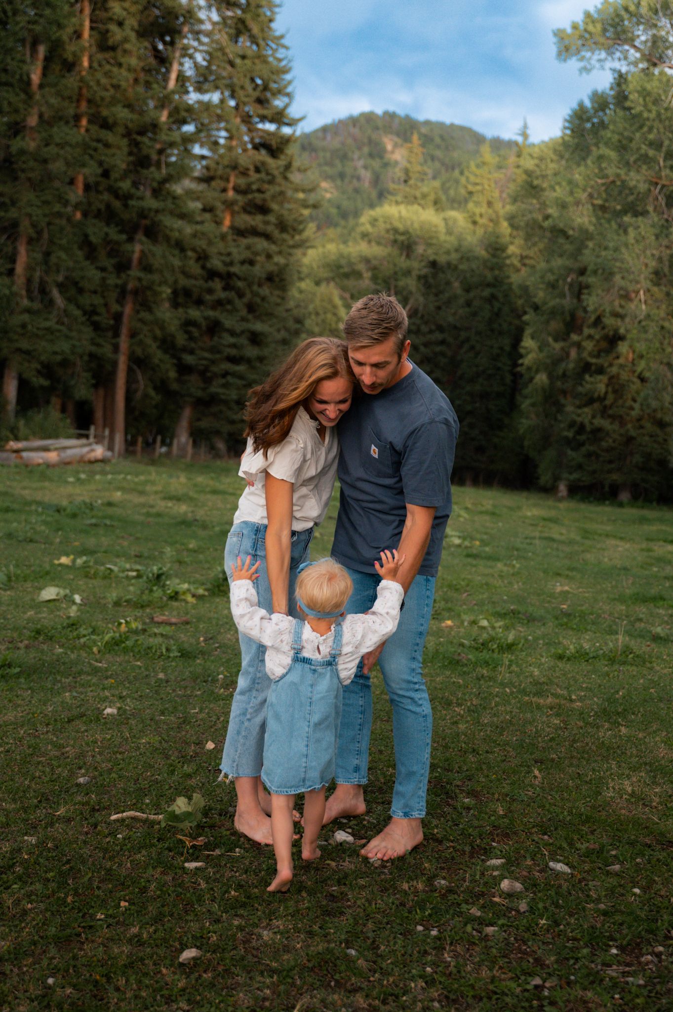 Young family with toddler embracing outdoors in a lush green meadow surrounded by pine trees.