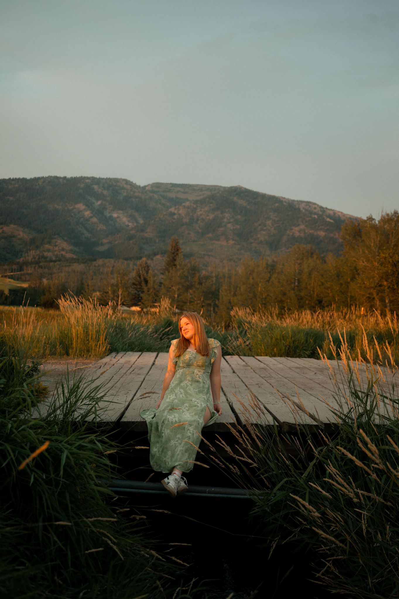 A woman in a green floral maxi dress sits on a wooden dock surrounded by tall reeds at sunset.