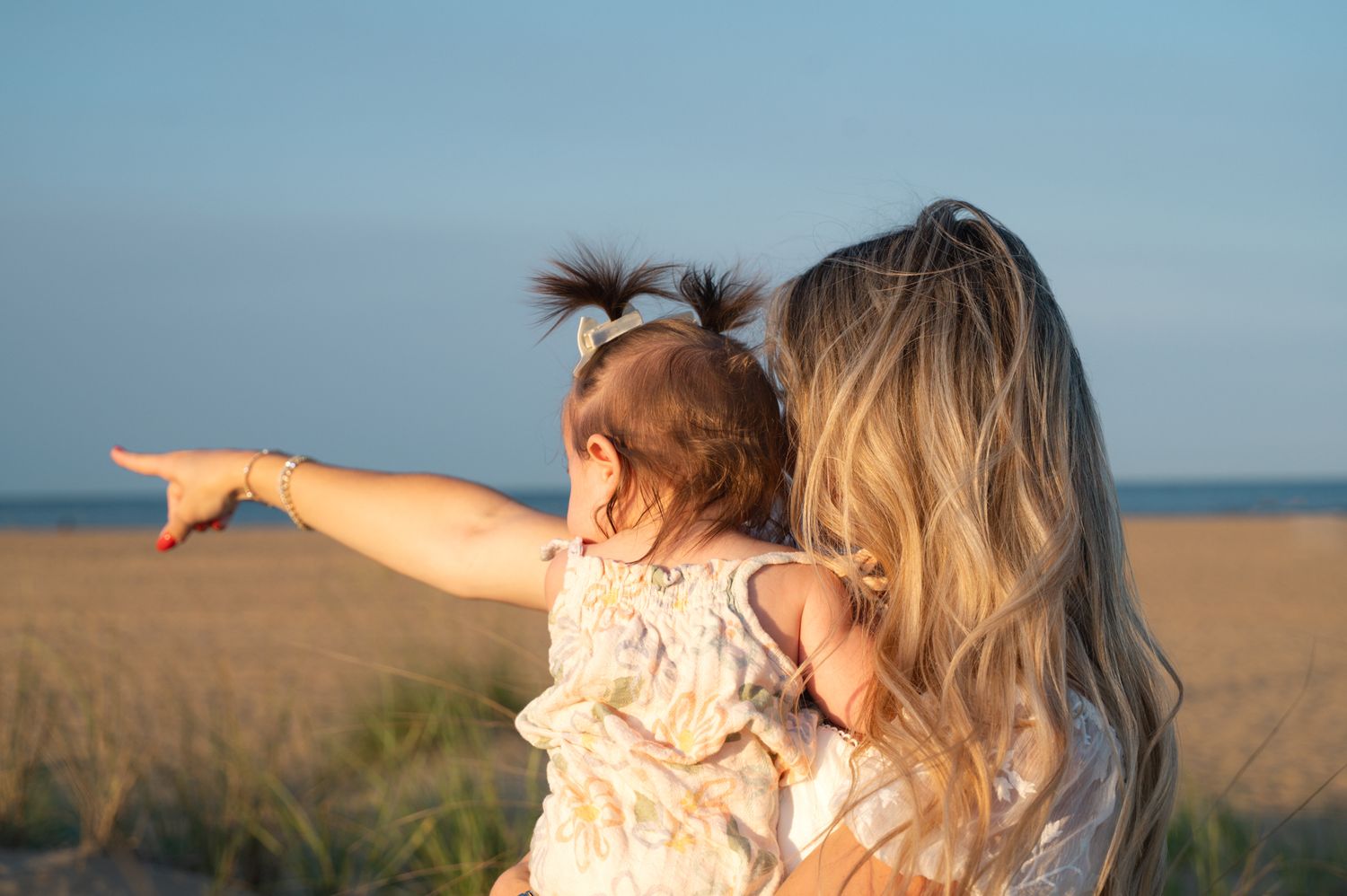 Mother holding toddler girl with pigtails pointing toward golden beach grass at sunset.